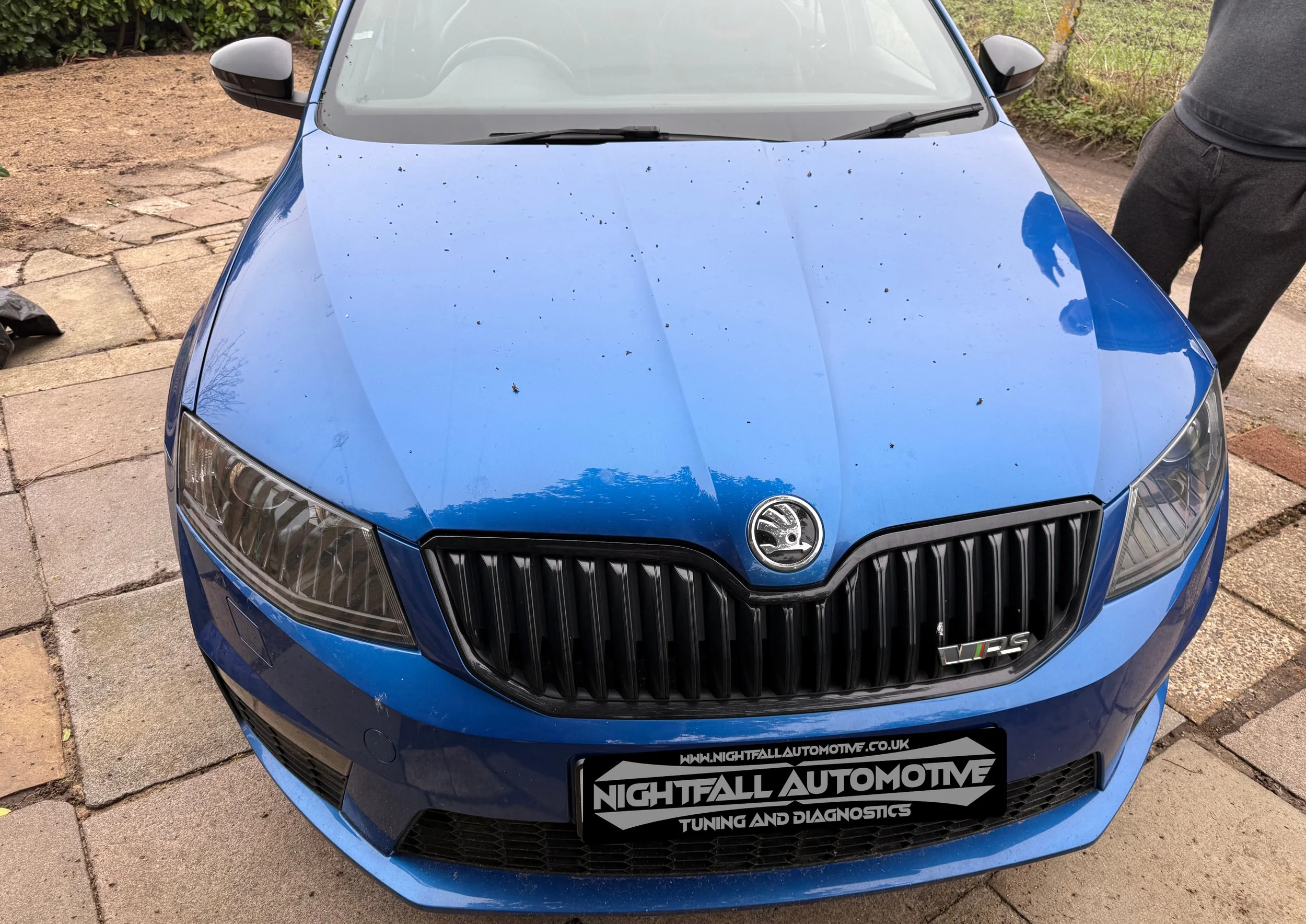 Front view of a blue hatchback car parked on a driveway, with dirt and leaves on its hood and a person standing next to it wearing gray sweatpants.