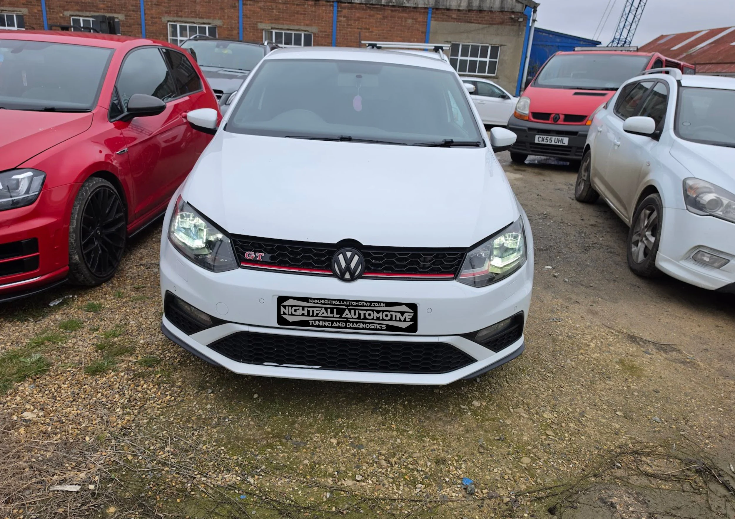 Front view of a white Volkswagen Golf GT car parked on a gravel lot, surrounded by other cars including a red car to the left and white cars on both sides, with a building in the background.