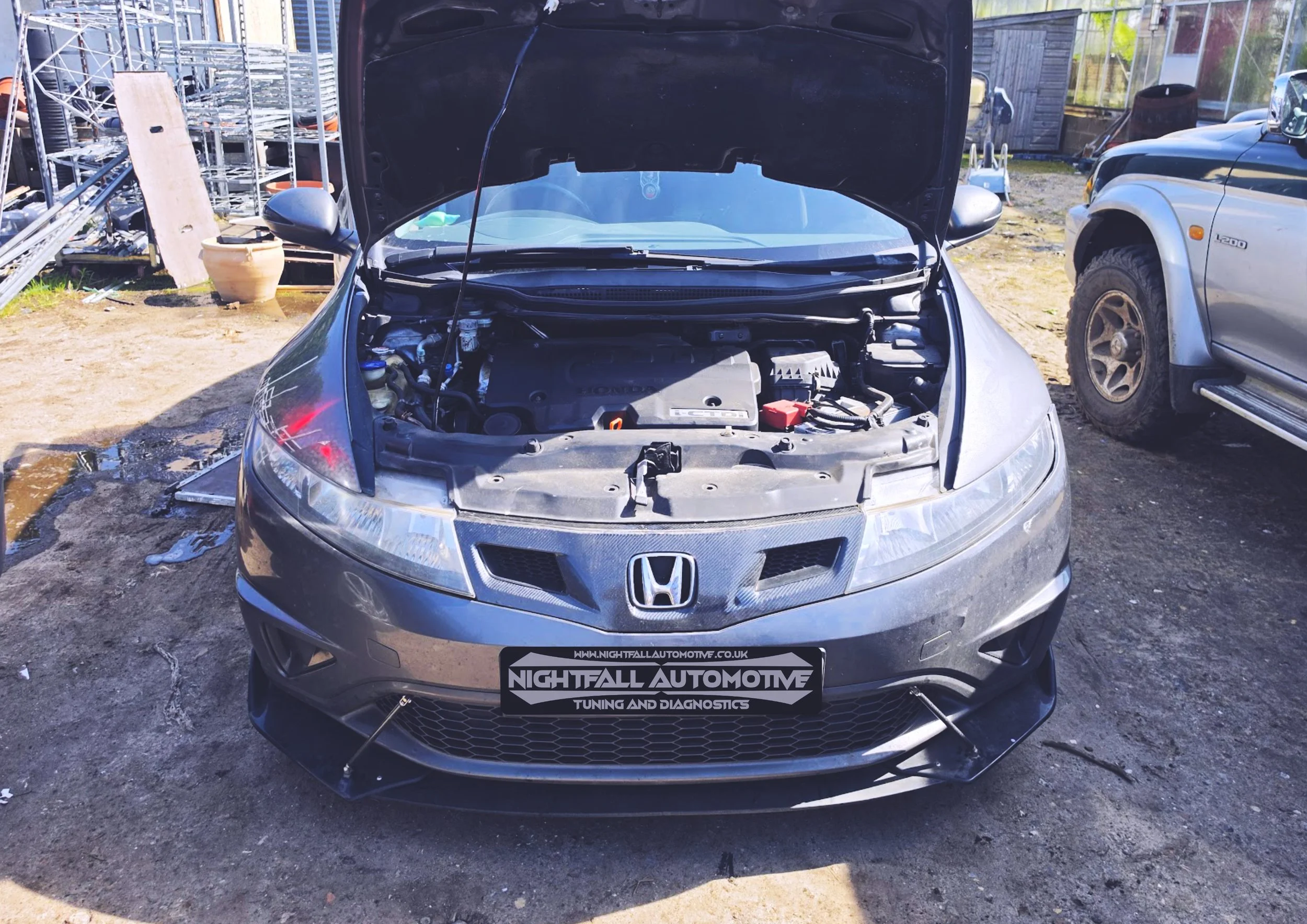 Front view of a gray Honda car with its hood open, situated outdoors in a mechanics or repair yard with various equipment and vehicles in the background. The front bumper has a black grille and a license plate with a website URL and branding for Nigh