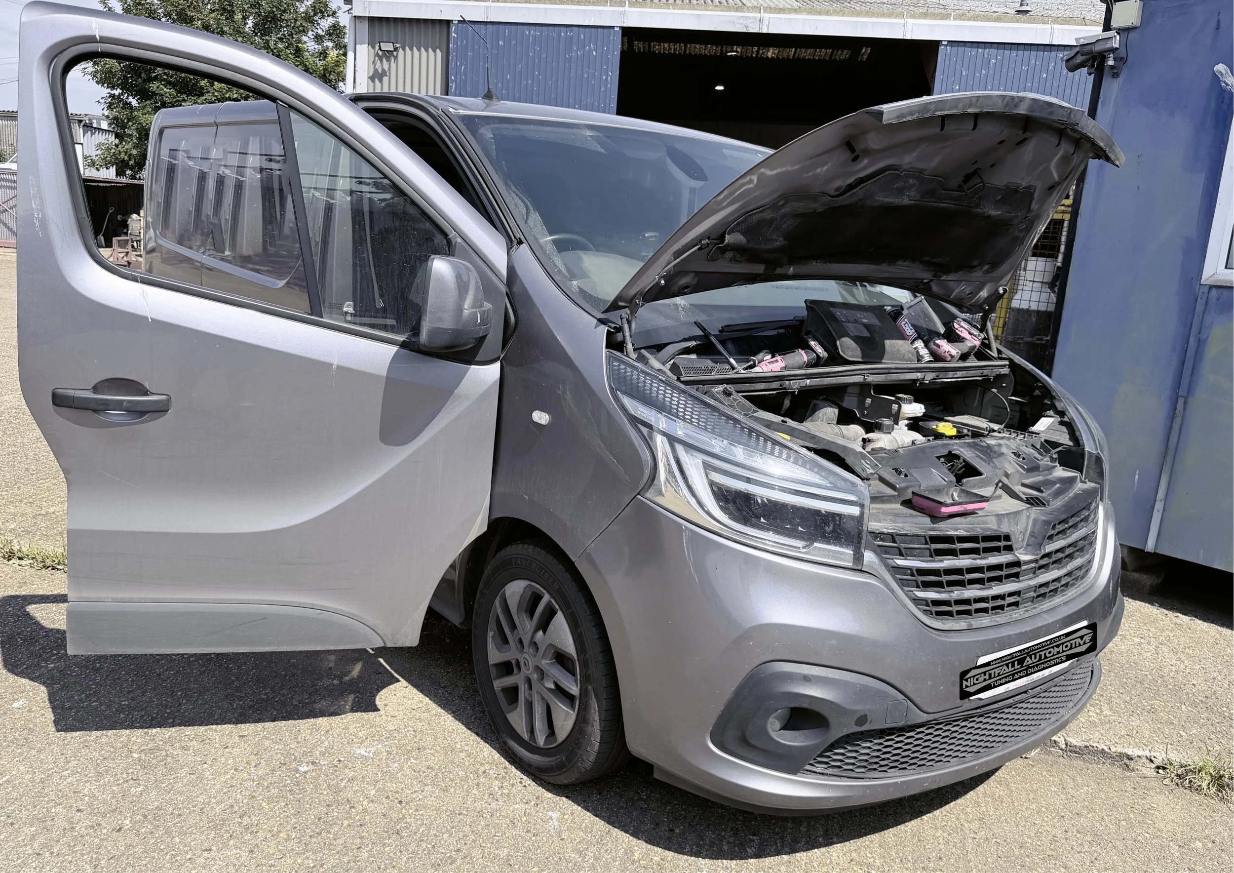 Silver car with its hood open revealing the engine, parked outside near a blue building with a garage door. Tuned by Nightfall Automotive