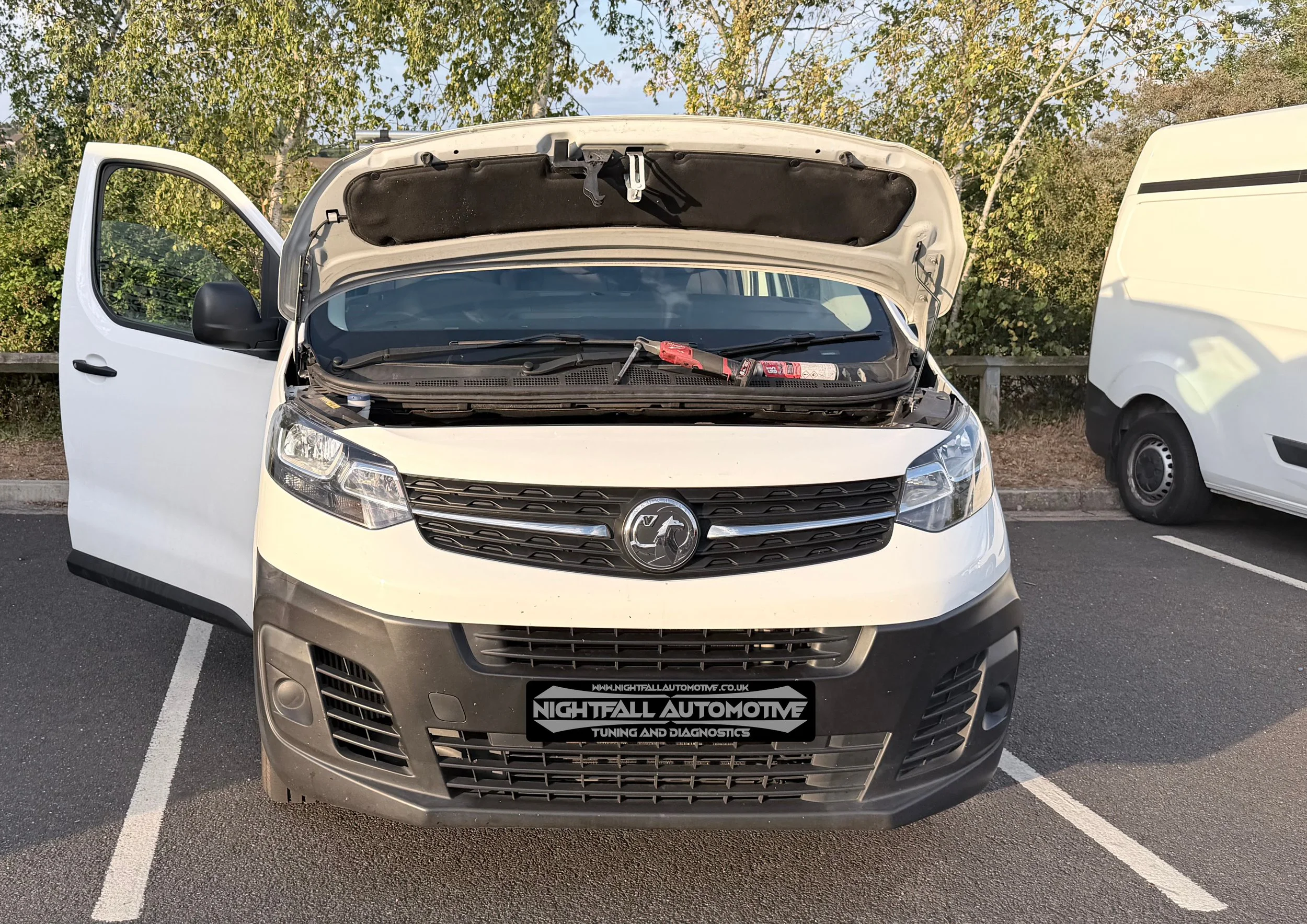 Front view of a white vehicle with its hood open, parked in a lot with trees in the background, and a sign that reads 'Nightfall Automotive Tuning and Diagnostics' on the front bumper. Tuned by Nightfall Automotive