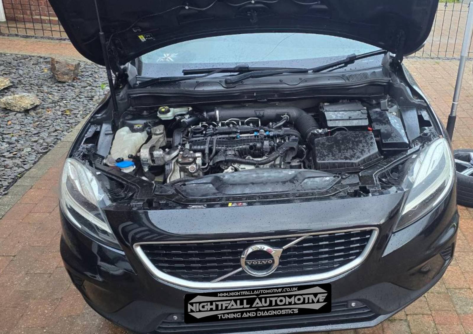 Front view of a black Volvo car with its hood open, showing the engine bay and various engine components, with a black grill and headlights visible, parked on a brick driveway.