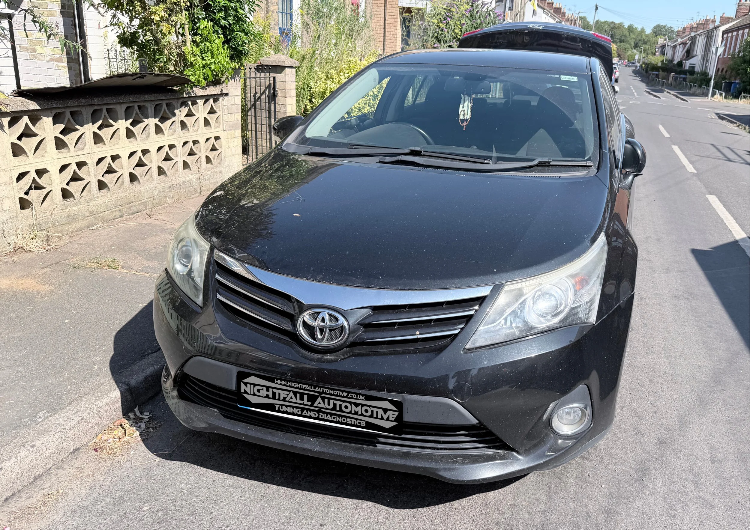 A black Toyota car parked on the street with a brick wall and greenery on the left and a residential street in the background. Tuned by Nightfall Automotive