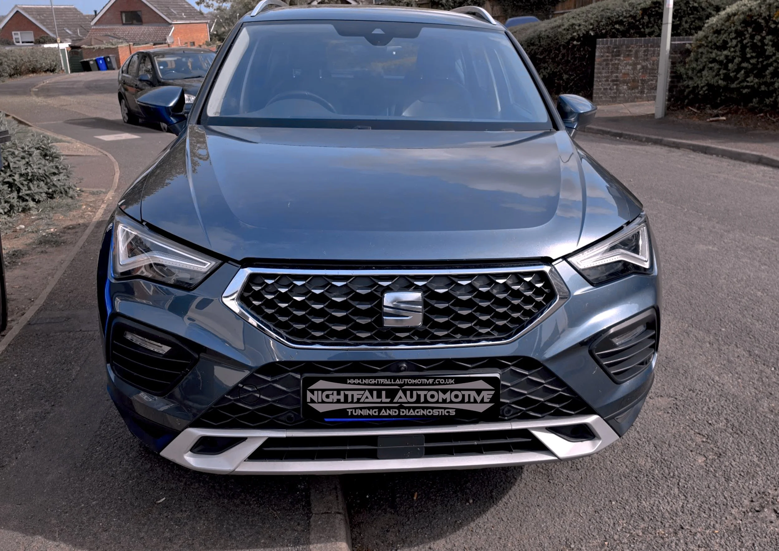 Front view of a metallic gray SEAT SUV parked on a residential street, with houses and another car in the background. The car features a black grille with a honeycomb pattern and LED headlights.