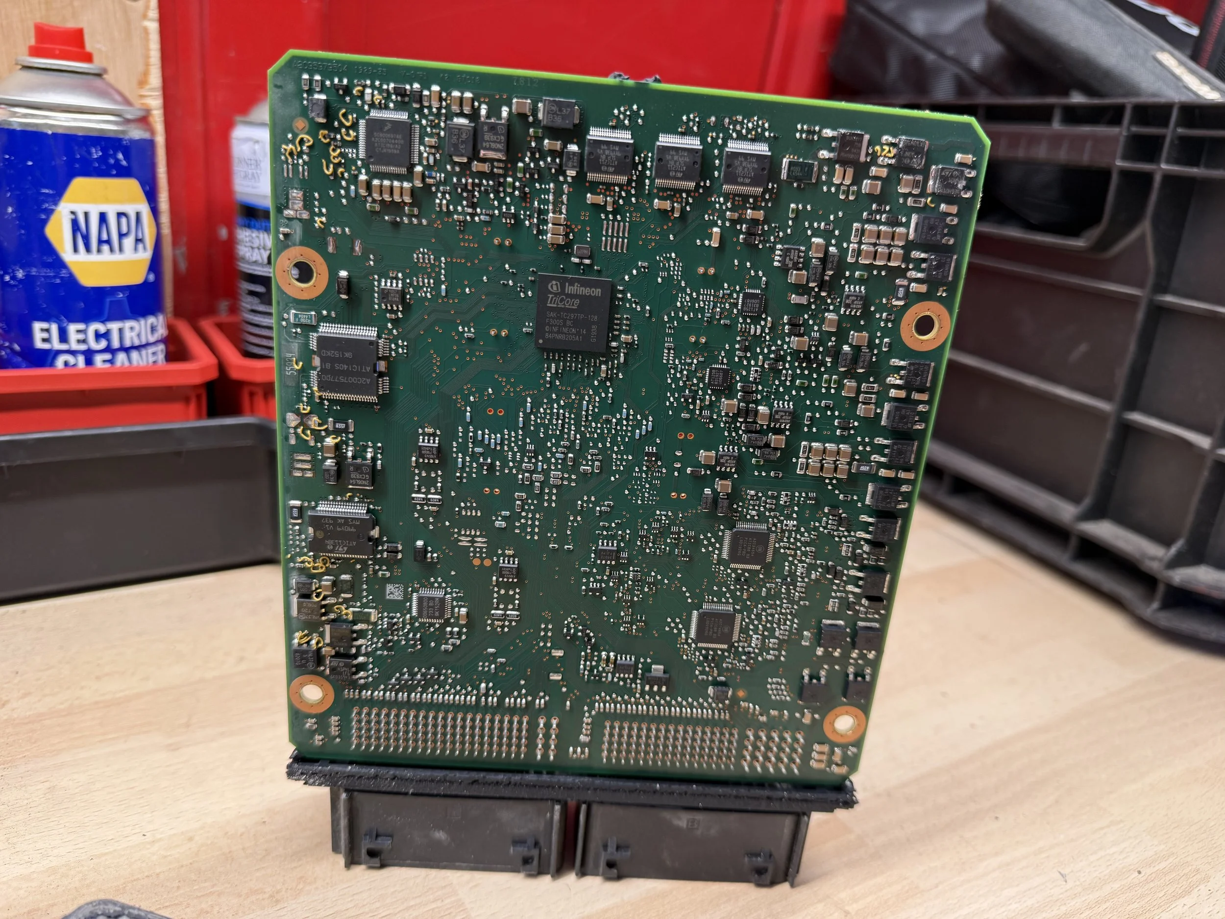 A green printed circuit board (PCB) with various electronic components and chips, propped up on a table in a workshop setting.