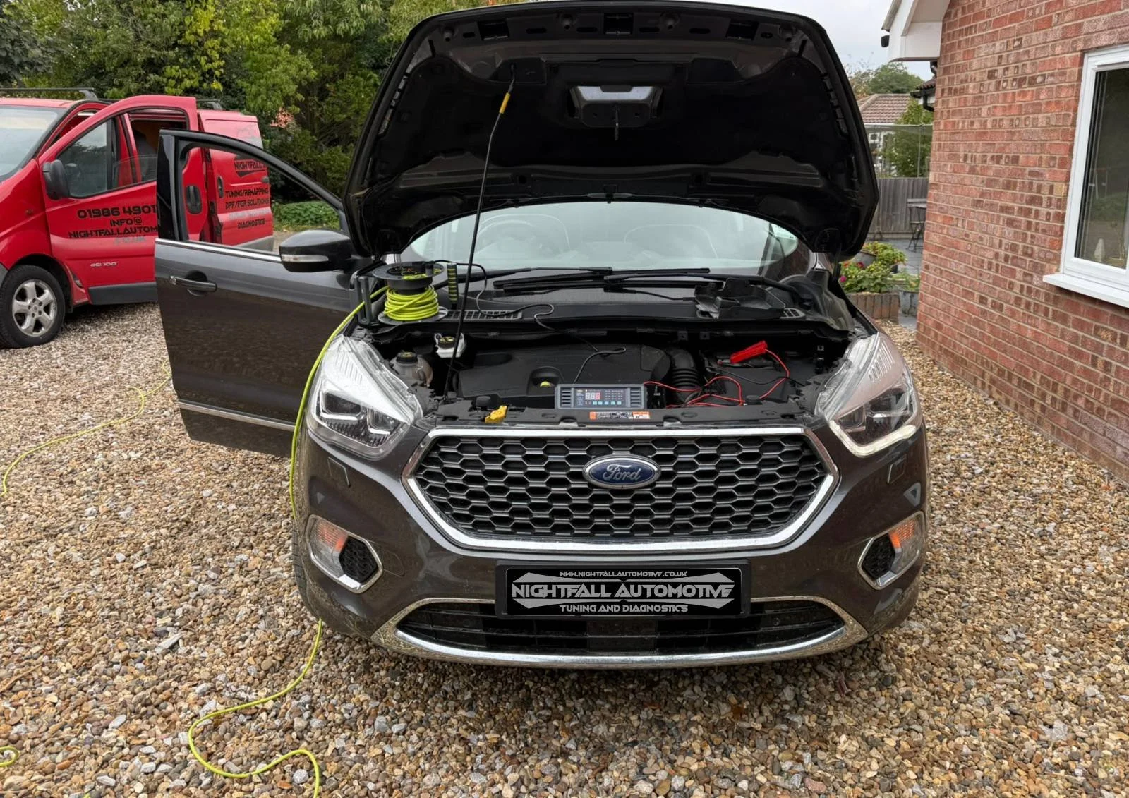 A black Ford vehicle with its hood open, undergoing tuning or diagnostics outdoors on a gravel driveway, with a red van in the background and a brick house on the side.