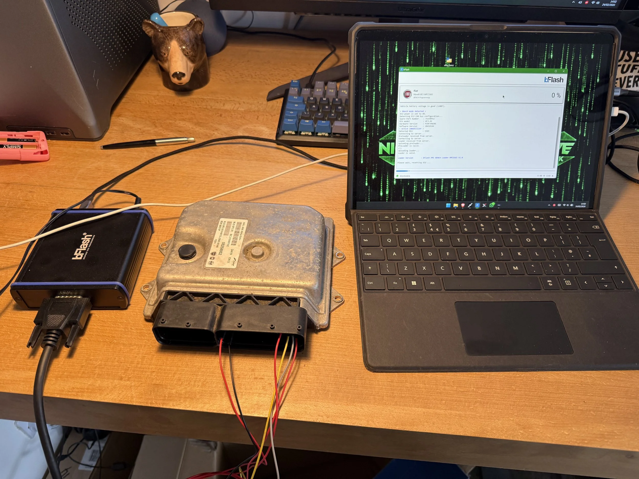 A workbench with a laptop displaying binary code and a flashing device connected via wires. An electronic module and a charger are on the table, with a pen, a small bear figurine, and a keyboard in the background.