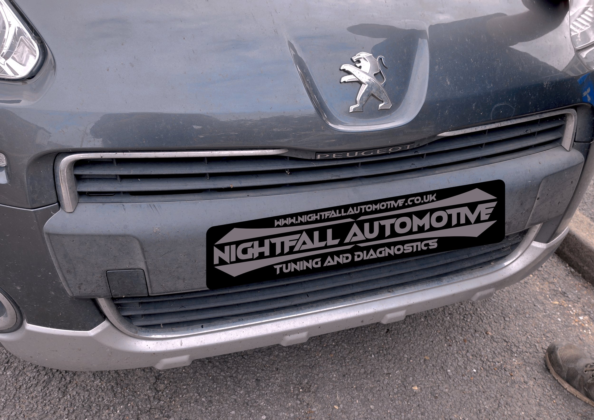 The front of a gray Peugeot vehicle with a lion emblem on the hood and a bumper sticker that reads 'Nightfall Automotive, Tuning and Diagnostics'.