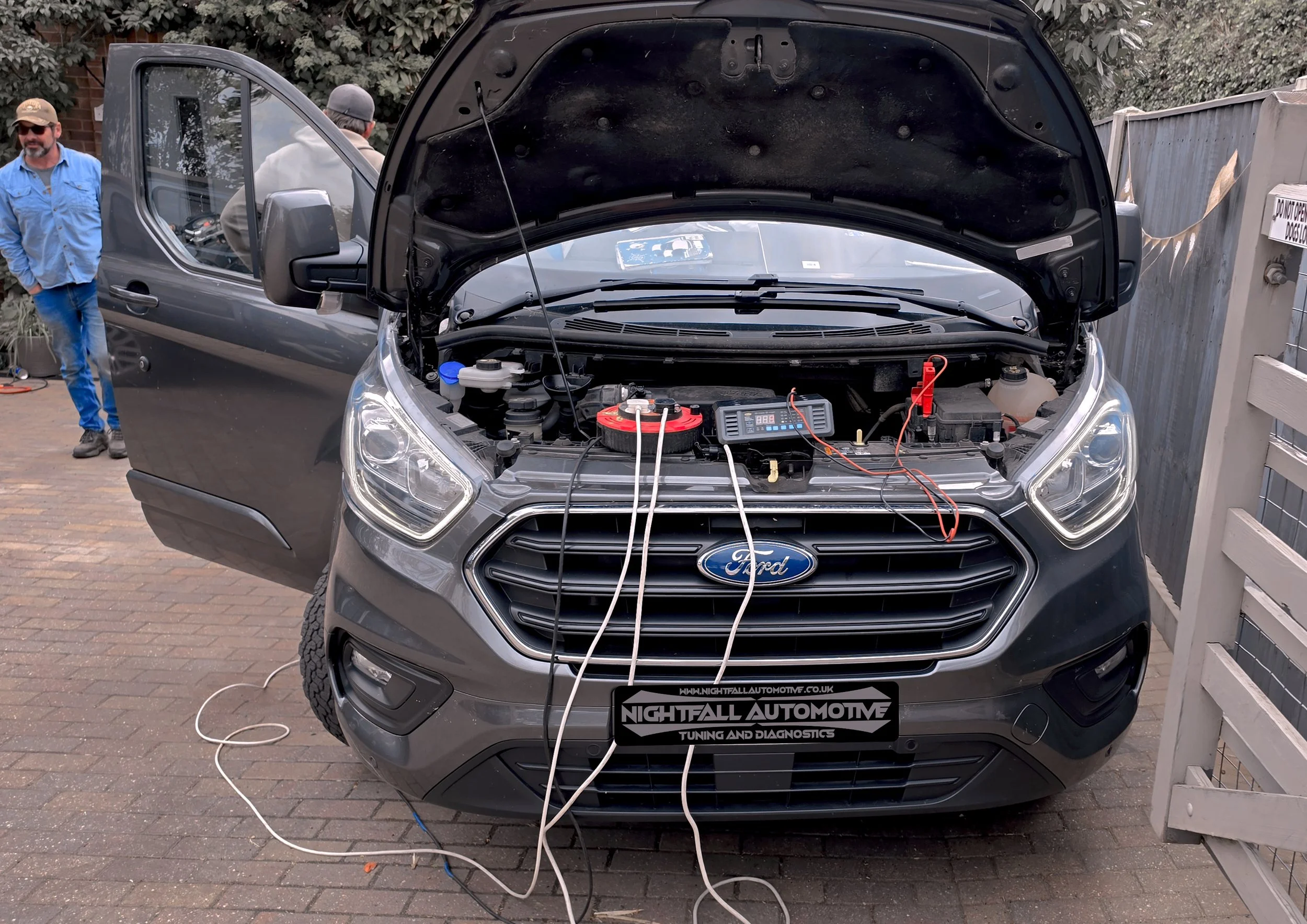 A black Ford vehicle with its hood open for maintenance or repair outdoors, with two men standing nearby, one wearing a blue shirt and sunglasses, the other wearing a gray hoodie and a gray cap. The engine diagnostics equipment and wires are visible 