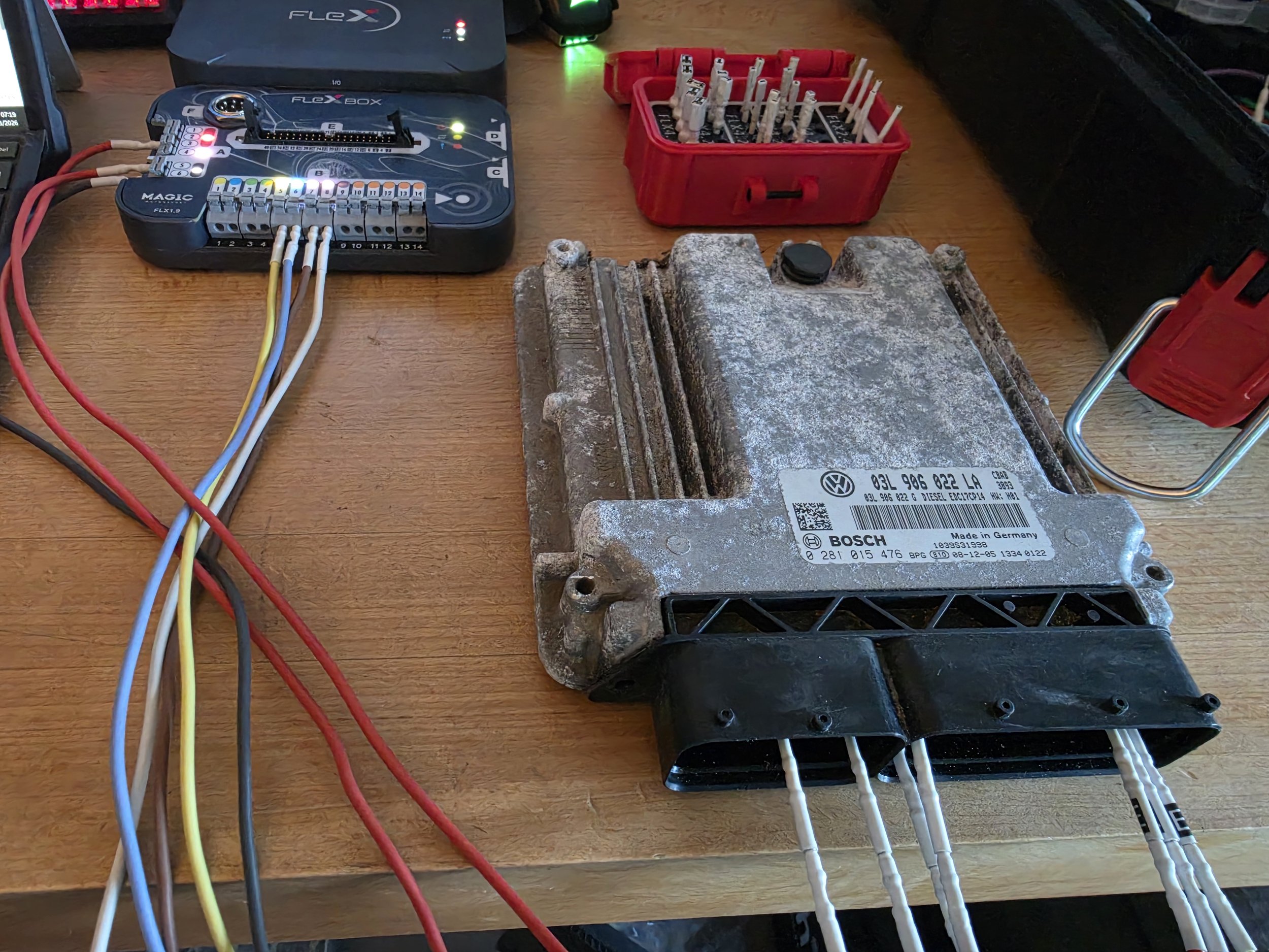 An automotive engine control unit (ECU) connected with multiple wires, placed on a wooden table with testing equipment and red plastic containers filled with electronic components.