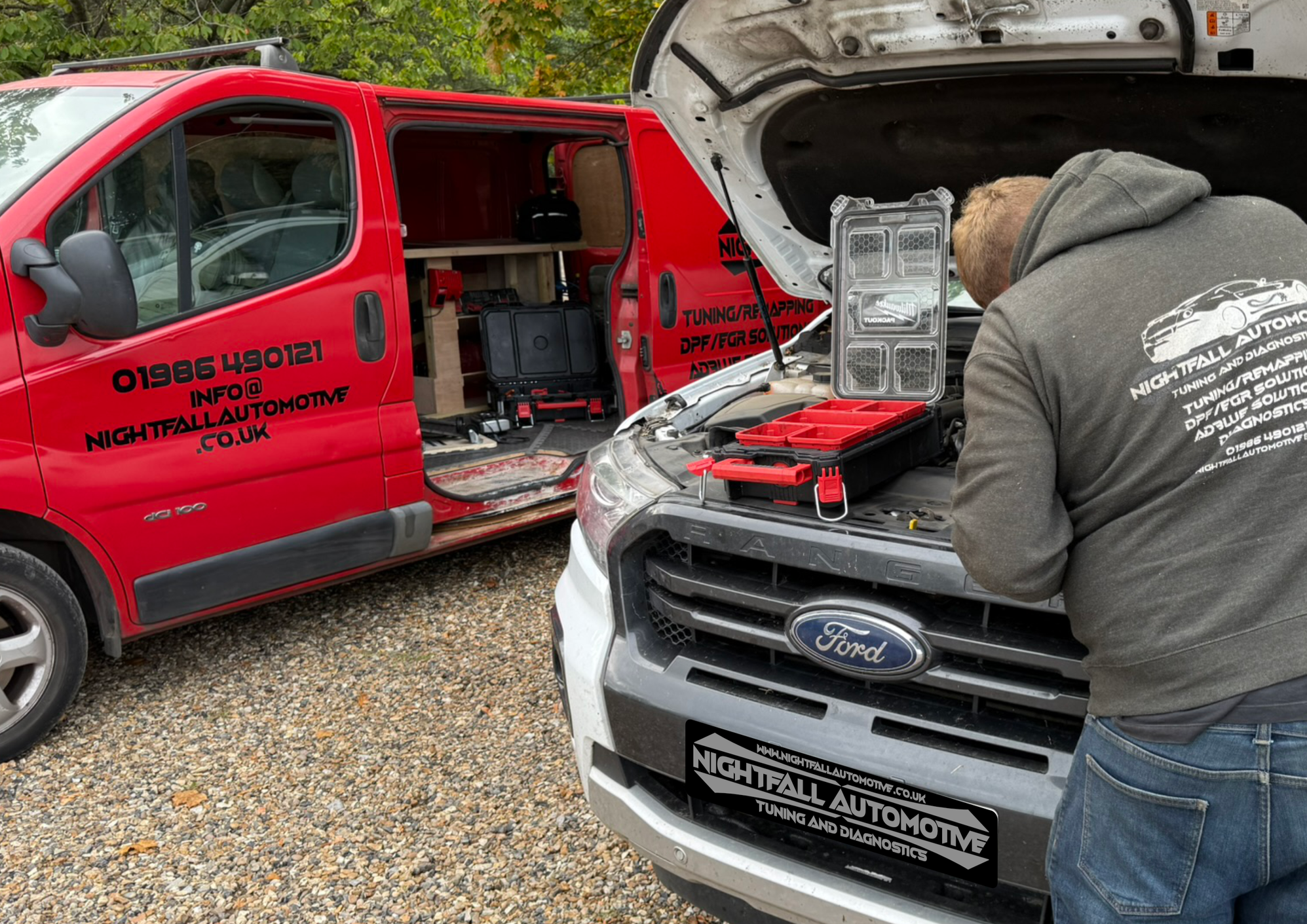 Two men working under the hood of a white Ford vehicle parked on a gravel surface, with a red van nearby. The men are wearing dark hoodies, and the van has text advertising 'Nightfall Automotive' with contact information and services, indicating an a
