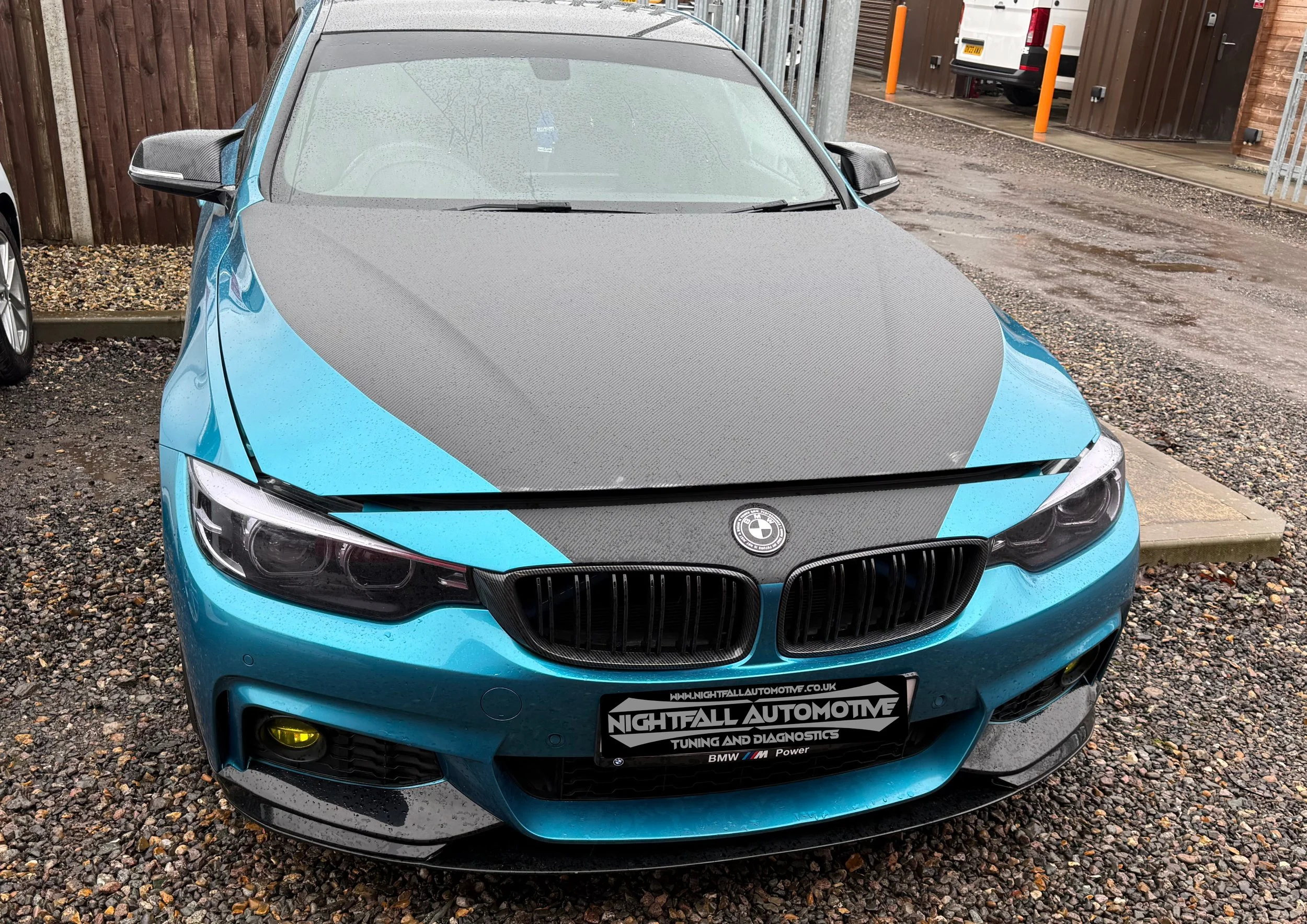 Blue BMW car parked outdoors on gravel, featuring a carbon fiber hood, daytime running lights, and Nightfall Automotive tuning and diagnostics decals.