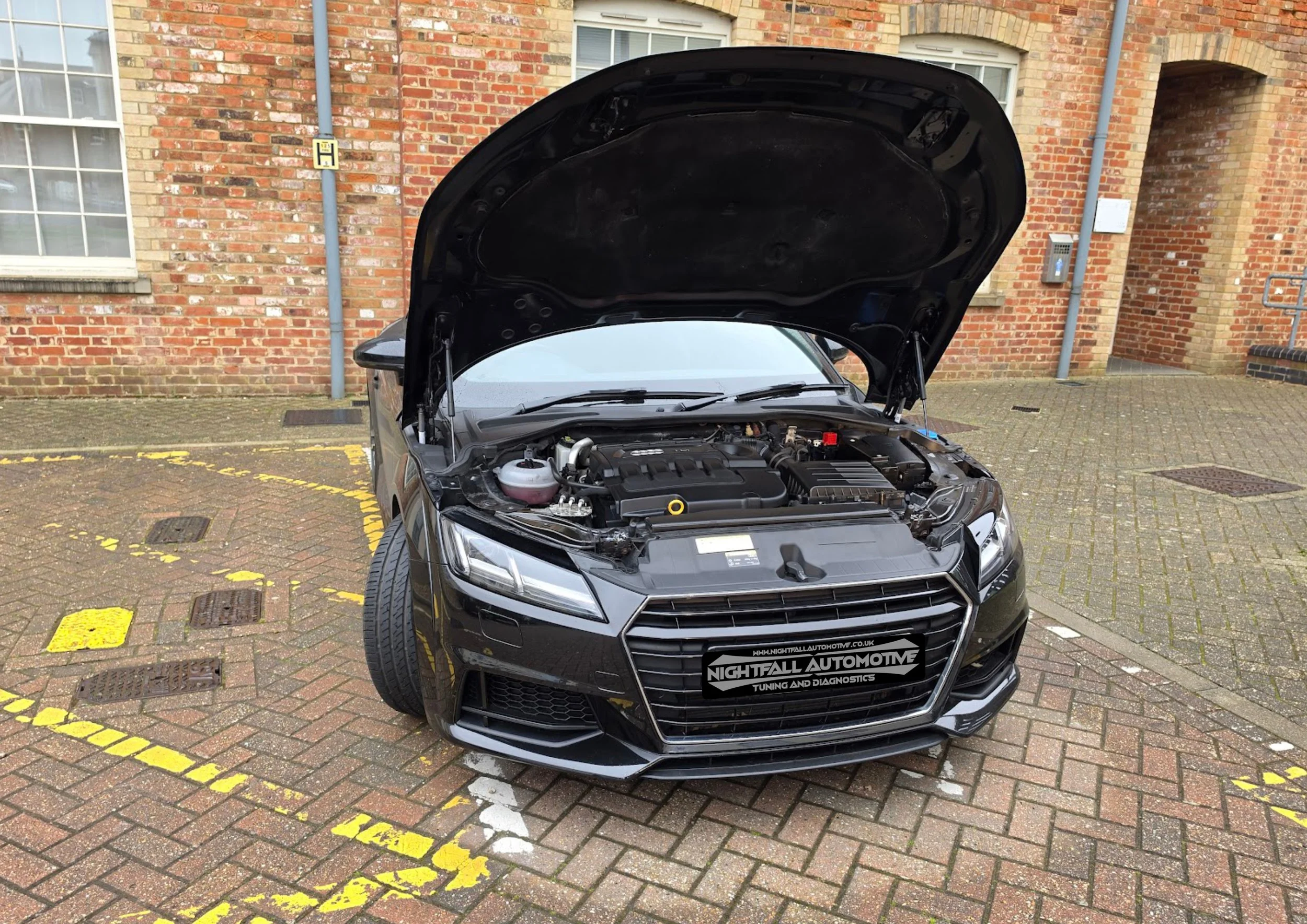 Black car with open hood parked on brick pavement in front of brick building, with yellow and white parking lines on the ground and a sign reading 'Nightfall Automotive Tuning and Diagnostics' on the front grille.