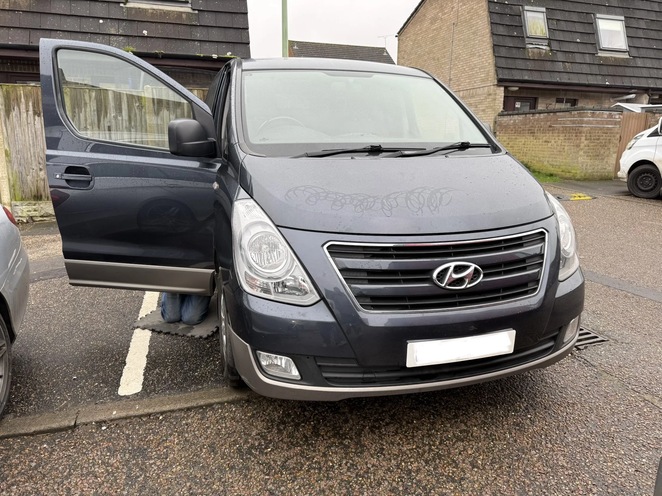 Dark gray Hyundai minivan parked in a parking lot with the driver's side door open. The van is wet from rain, and there is a pair of shoes visible under the open door. DPF solution by Nightfall Automotive
