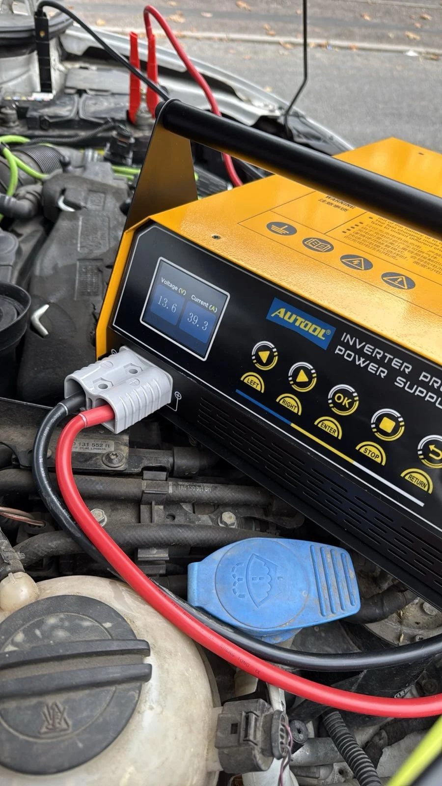 A yellow AUTOOL inverter power supply device connected to a car battery. The device displays voltage of 13.6V and current of 39.3A, with several control buttons underneath. The car's engine bay is visible, with various wires, hoses, and components.