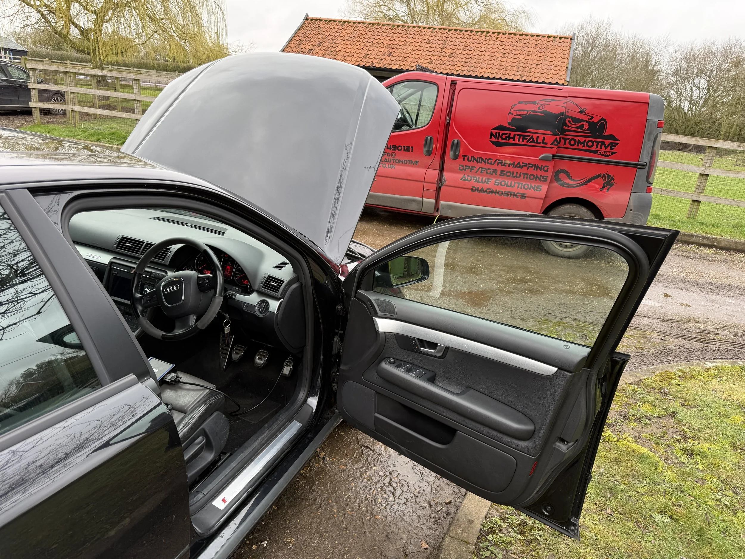 A black Audi car with its driver's side door open, showing the interior with steering wheel, dashboard, and pedals, parked outdoors near a red work van with graphics and text for Nightfall Automotive, with a hood open on the car and a building with a