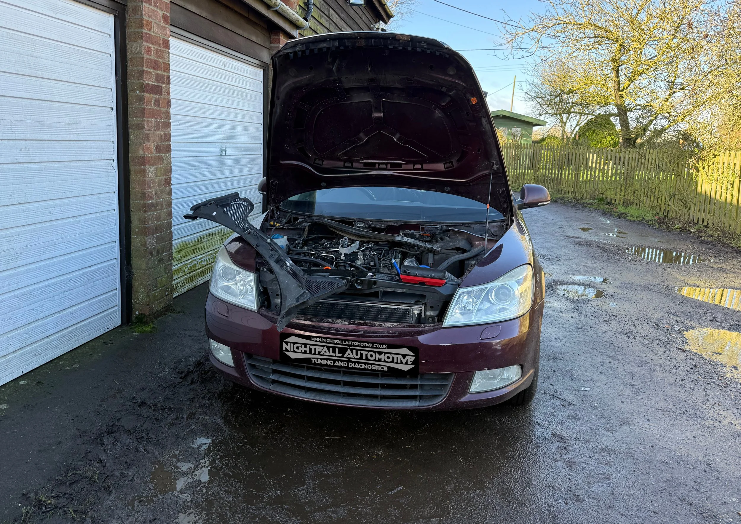 A maroon car parked outside with its hood open, showing the engine. The car is in a driveway next to a garage with a white door and a brick wall. The license plate has a logo for Nightfall Automotive. The ground is wet and there are puddles nearby.