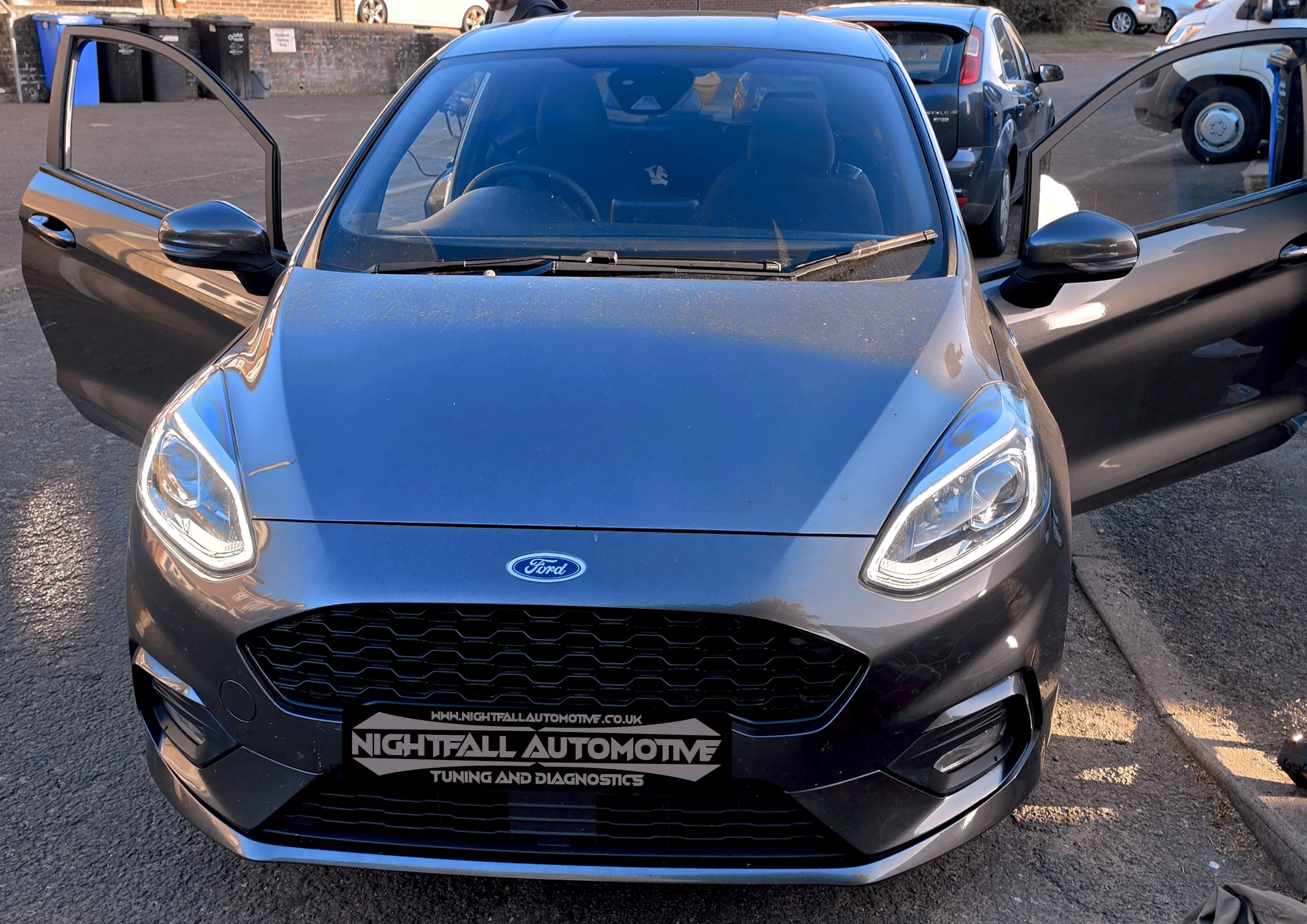 Front view of a metallic gray Ford car with both front doors open, showing the interior, parked in an outdoor parking lot with other vehicles and waste bins in the background.