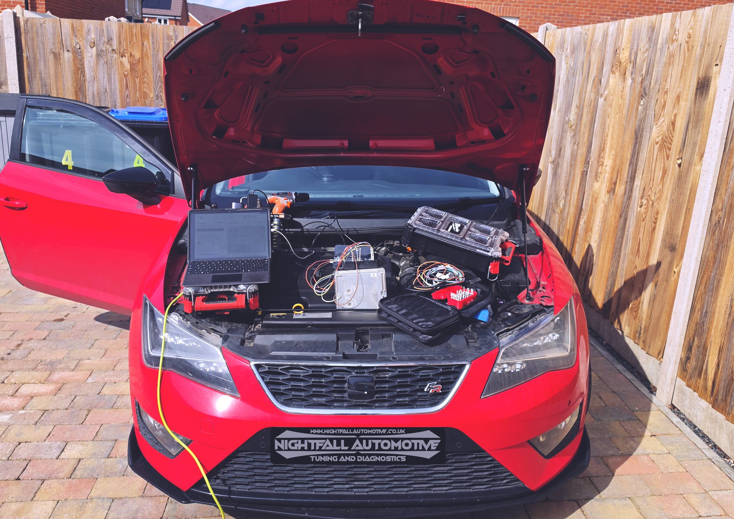 Red car with its hood open, displaying the workbench with electronic diagnostic tools and equipment for tuning and diagnostics, parked outdoors with a wooden fence in the background.
