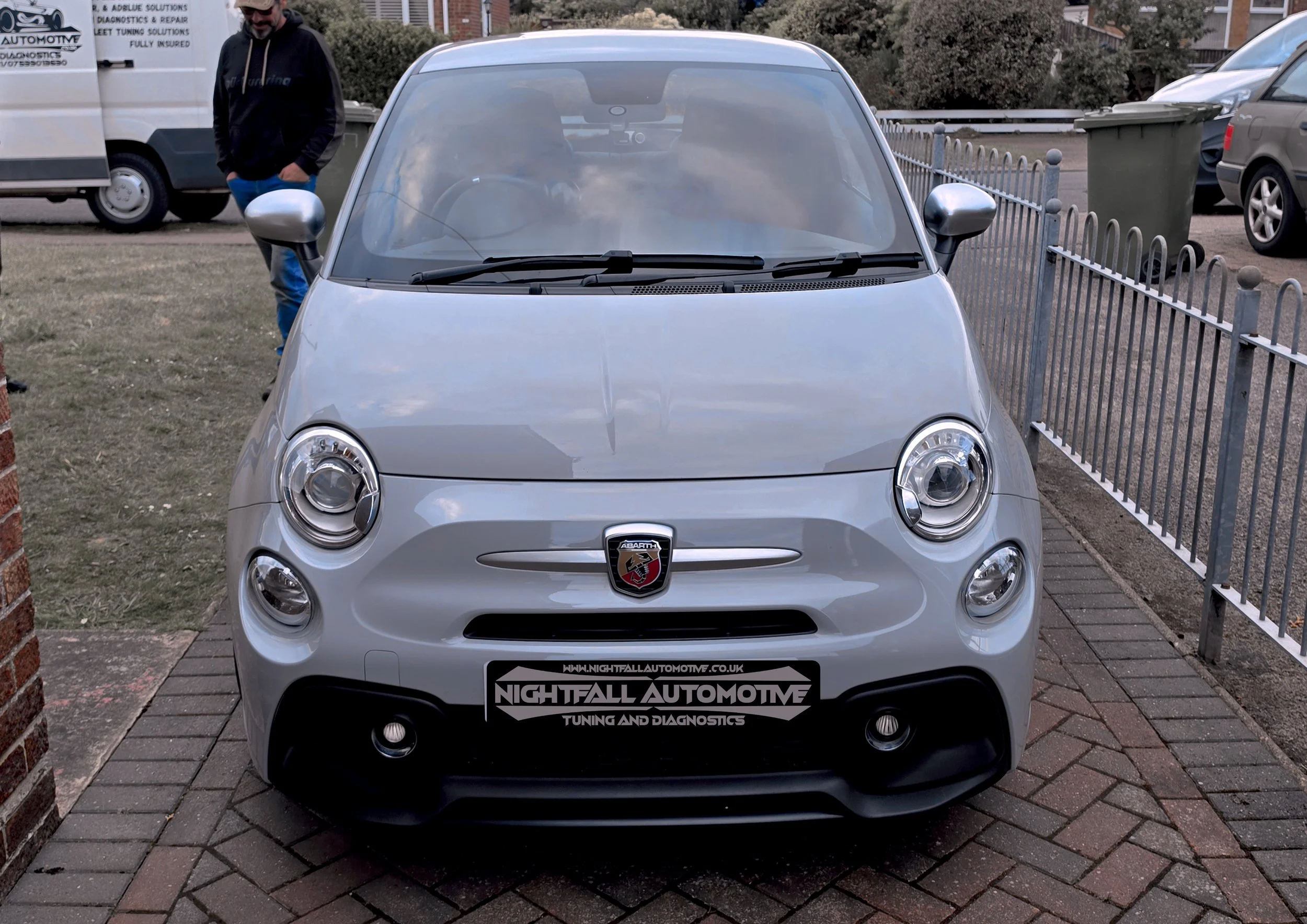 Front view of a silver Fiat Abarth car parked on a brick driveway. The car has a black front bumper, silver side mirrors, and the Abarth logo on the front grille. There is a banner on the front of the car with the text 'Nightfall Automotive, Tuning a