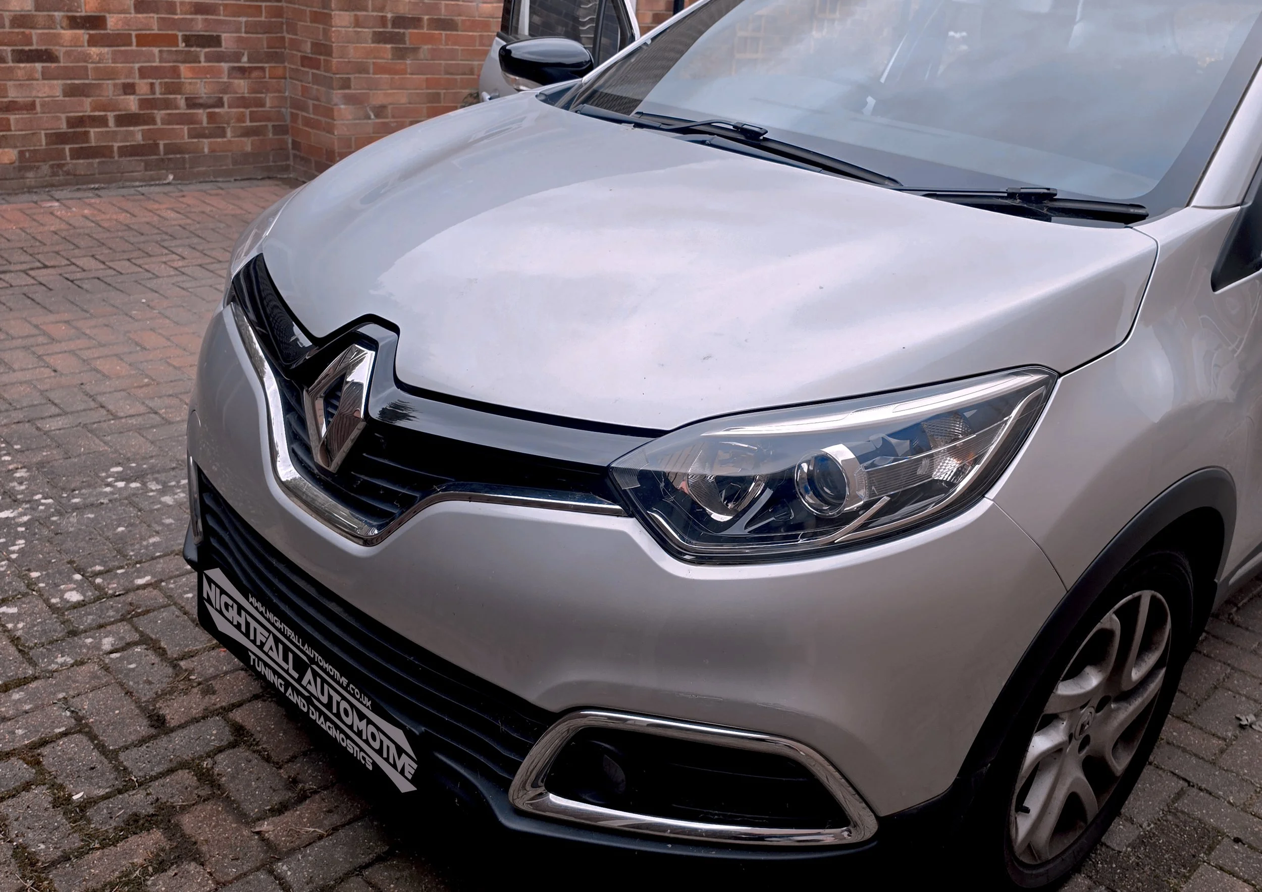 Close-up of a silver car parked on a brick-paved surface, with a brick wall in the background. The front of the car, including the headlight and grille, is visible. The car has a black license plate with white text.