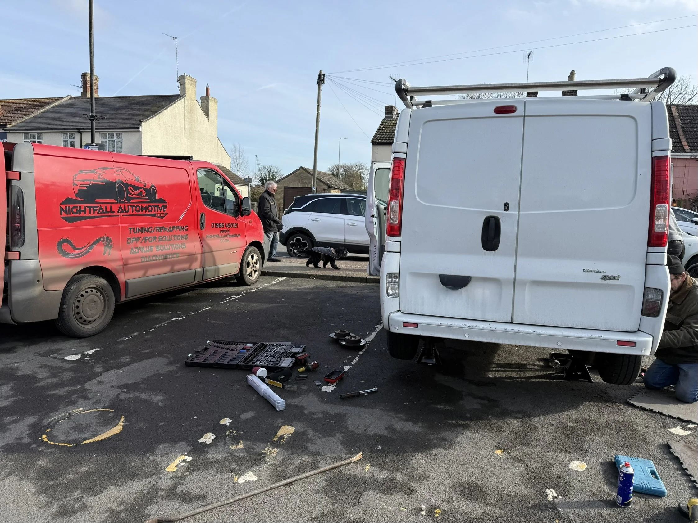 Automotive repair scene in a parking lot with a red service van labeled 'Nightfall Automotive' and a white van being worked on, tools and equipment scattered on the ground, a person kneeling repairing, and another man walking a black dog.