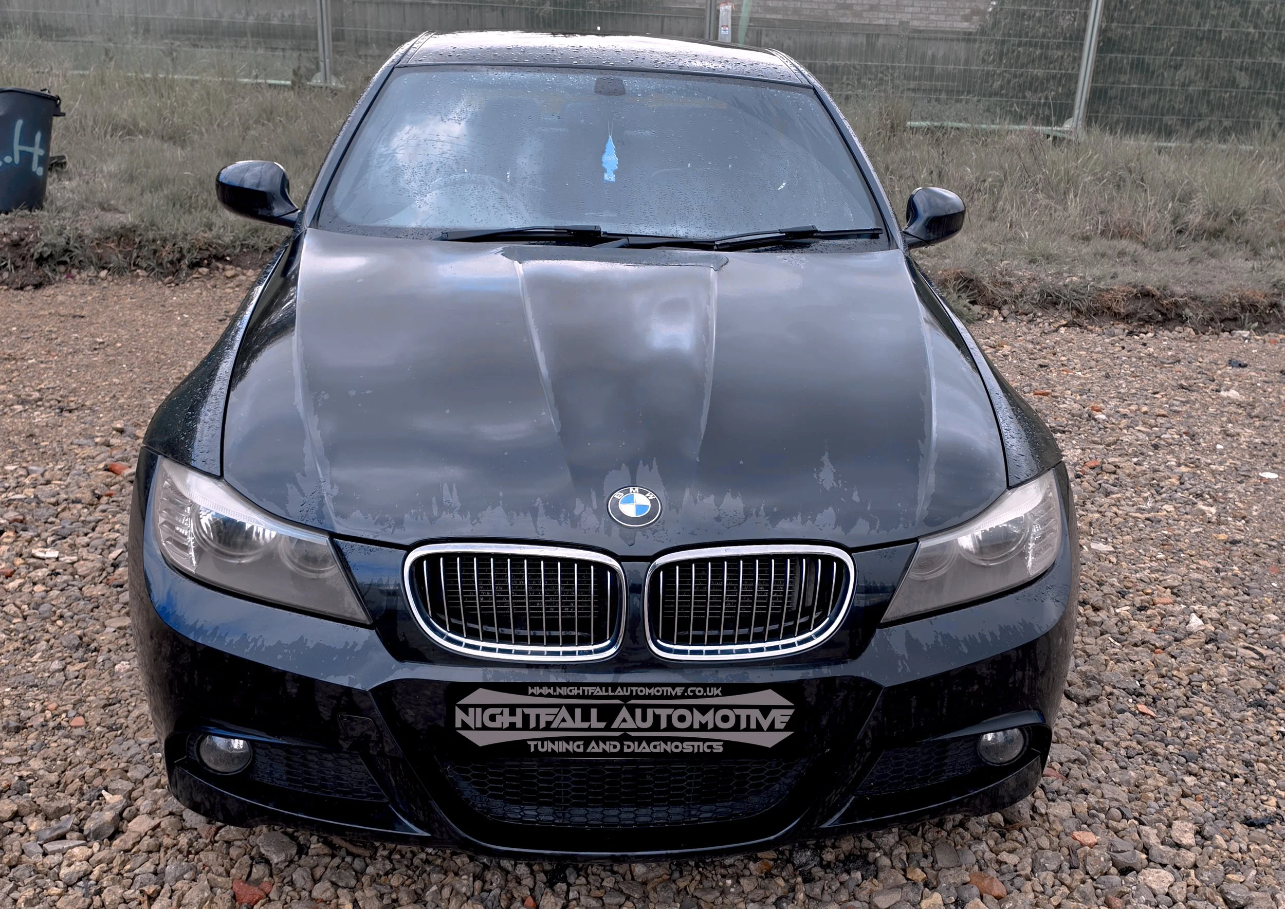 A black BMW car with water droplets on its surface, parked on a gravel lot. There is a fence and some grass in the background. The front grille displays the BMW logo and a sticker at the bottom reads "Nightfall Automotive Tuning and Diagnostics."