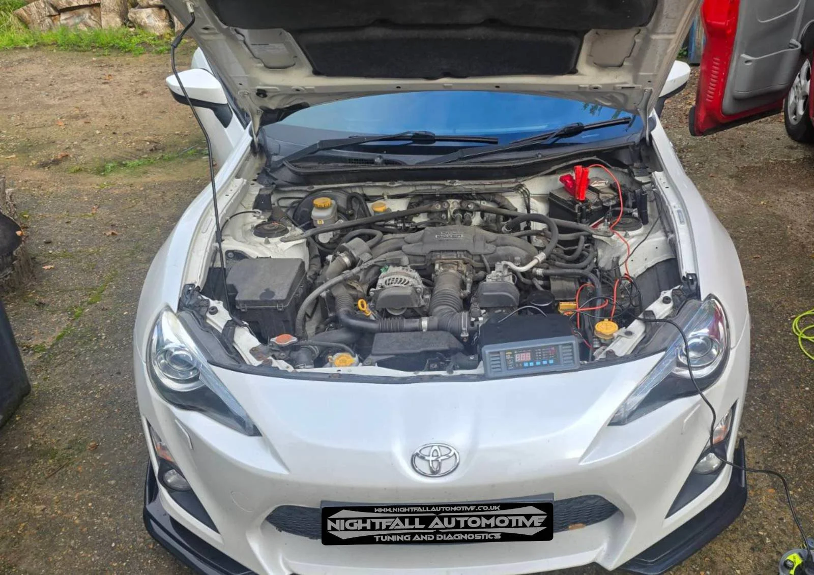 A white Toyota sports car with its hood open, revealing the engine bay. There is diagnostic equipment connected to the engine, and the garage has an outdoor, gravel area with some tools and parts around.
