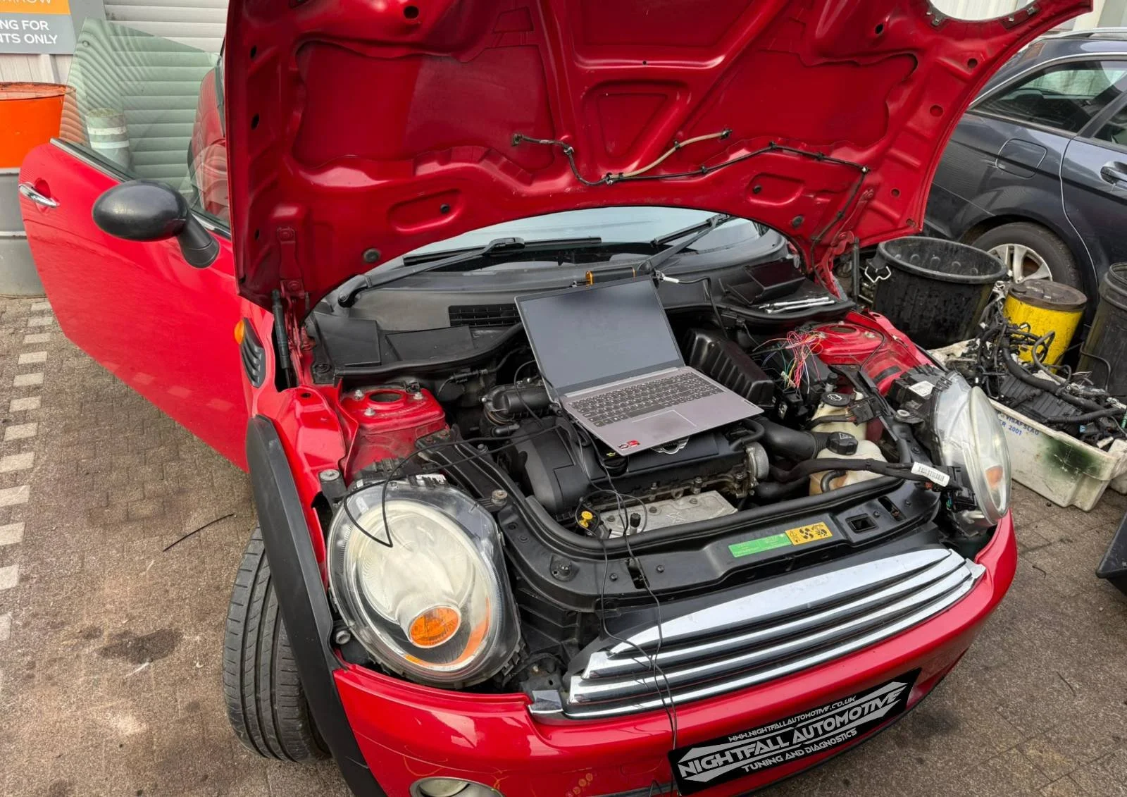 Open hood of a red Mini Cooper car with a laptop on the engine, surrounded by workshop tools and equipment.