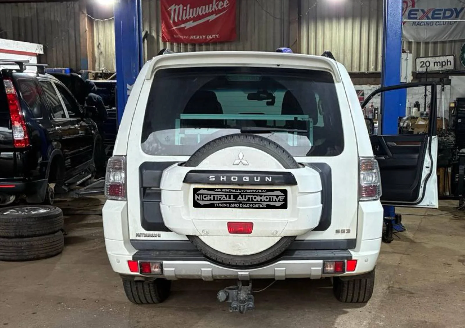 White Mitsubishi Shogun SUV inside an automotive repair shop, with a spare tire mounted on the rear door and a towing hitch below. The shop has tools, tires, and equipment visible around, with signs and banners on the walls.
