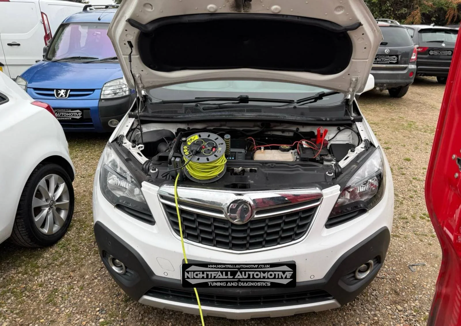 White car with open hood showing engine bay and diagnostic equipment connected, parked among other vehicles at a dealership lot.