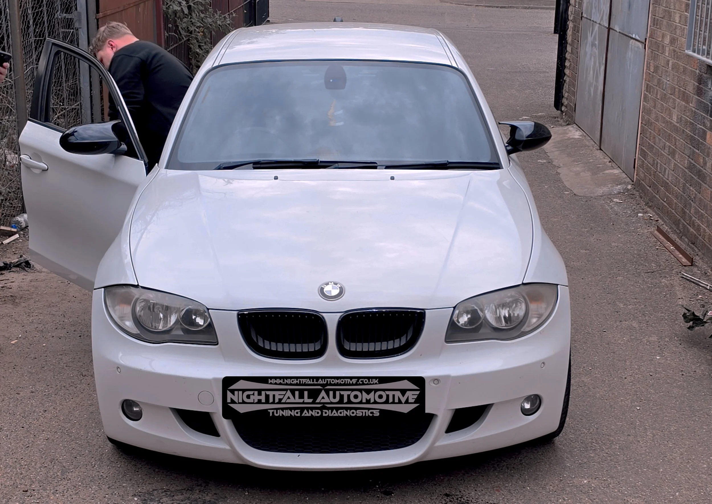 White BMW car parked with front view showing the logo on the hood, with a person in black clothing leaning into the driver's side door. tuned by Nightfall Automotive