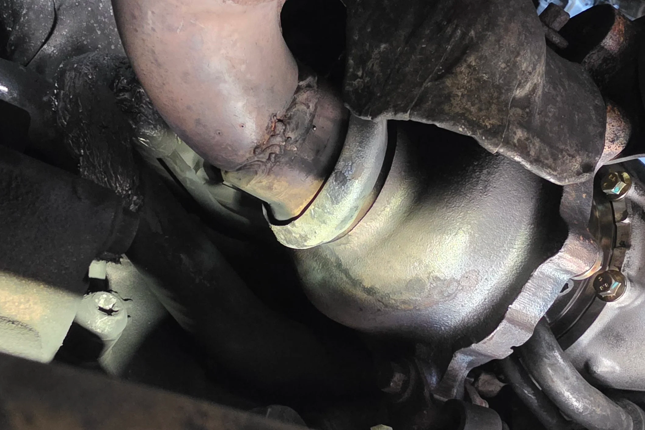 Close-up view of a rusty metallic engine component with welded joints and connected pipes, likely part of a vehicle's exhaust system. Nightfall Automotive tuning and diagnostics