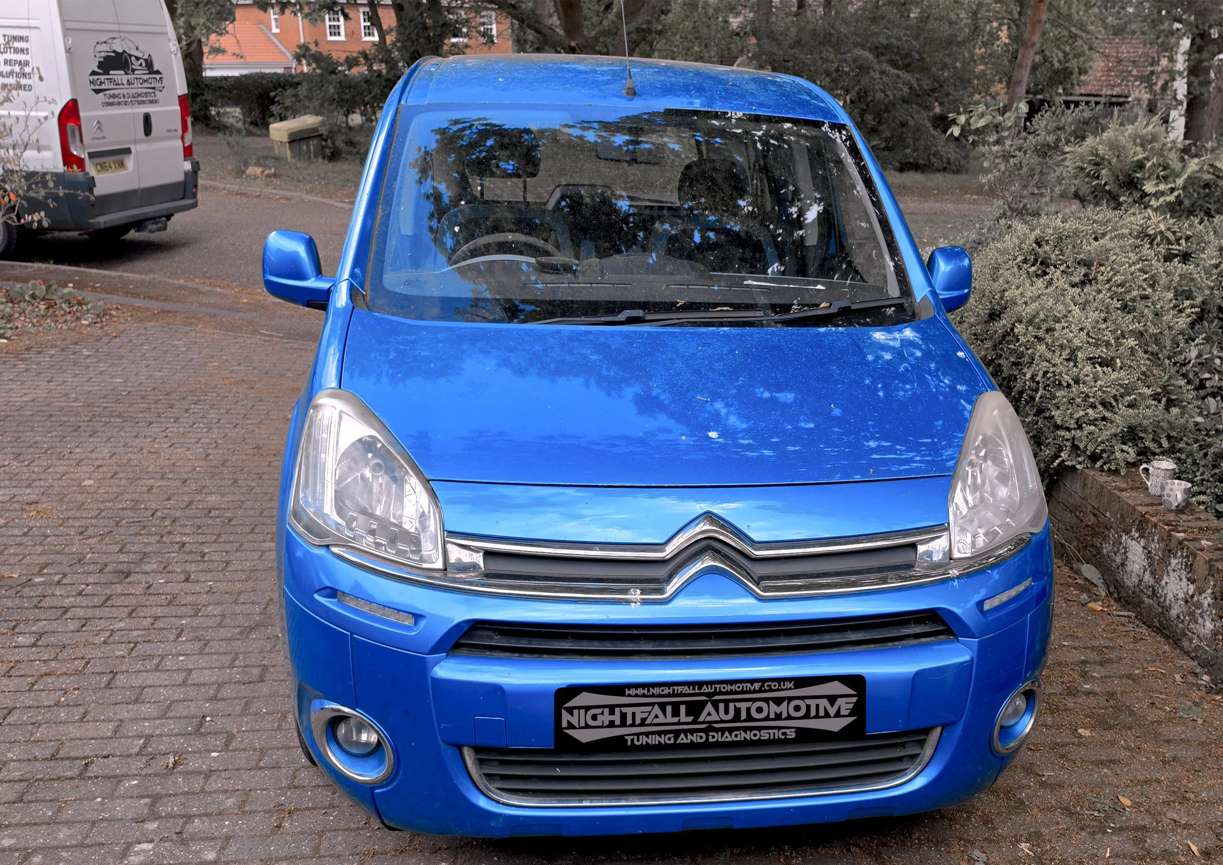 Front view of a shiny blue compact car parked on a brick driveway, with another van and greenery in the background.