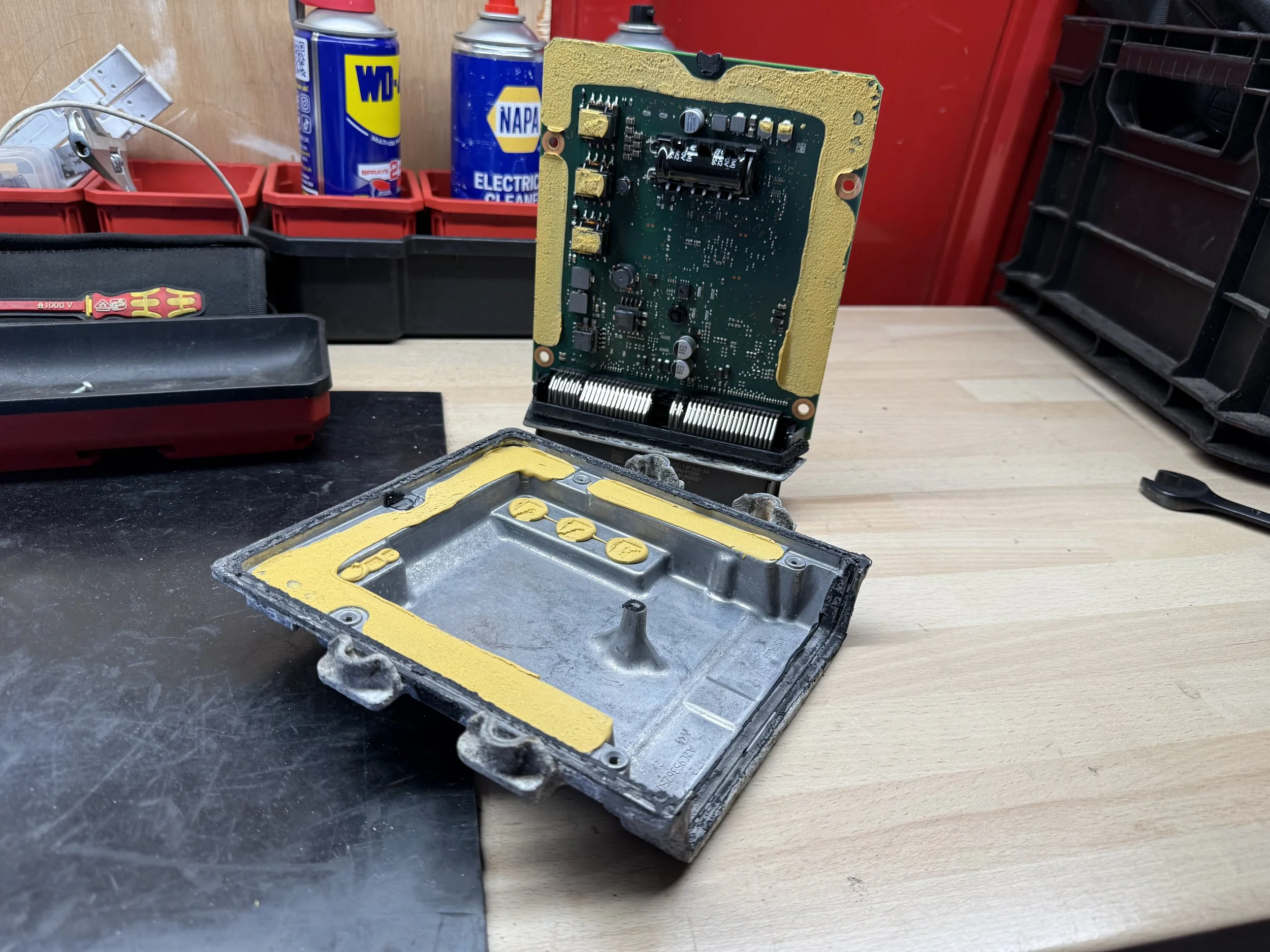 Disassembled electronic device with a circuit board and metal casing on a workbench, with tools and shelves in the background.