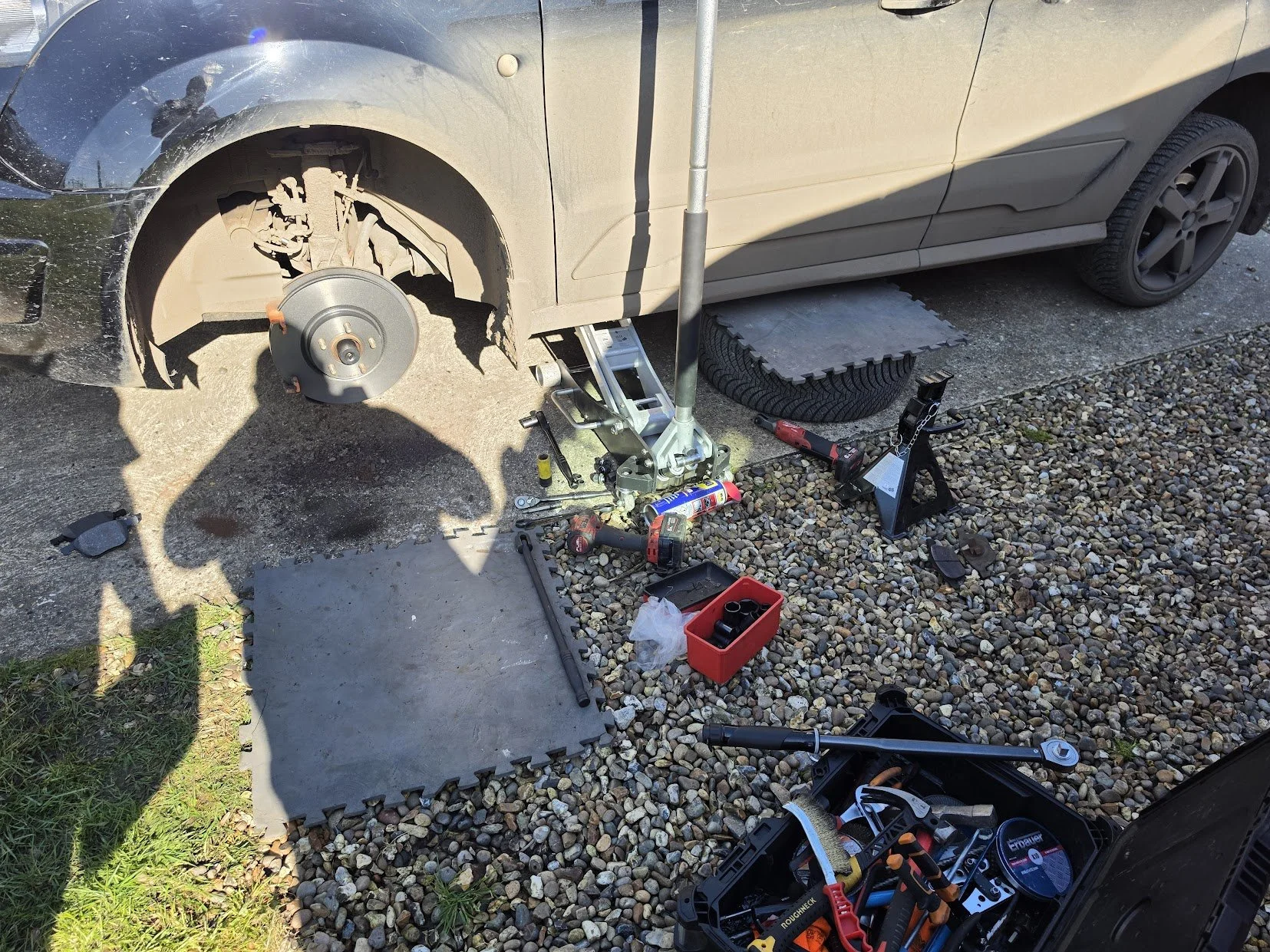 A car with the front wheel removed, exposing the brake disc. The car is elevated using a hydraulic jack, and various tools and parts are scattered around on the ground, including a tire, a creeper, and a toolbox, suggesting the car is being repaired 