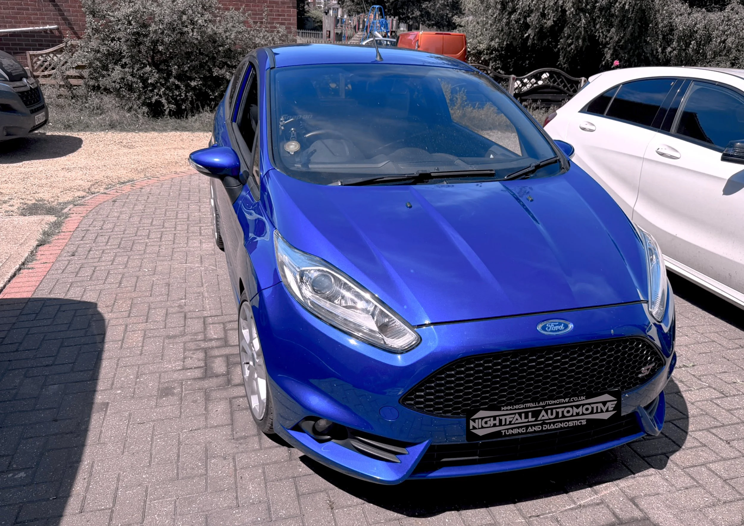A blue Ford car parked in a lot next to a white car, with some bushes, a brick wall, and other cars in the background.
