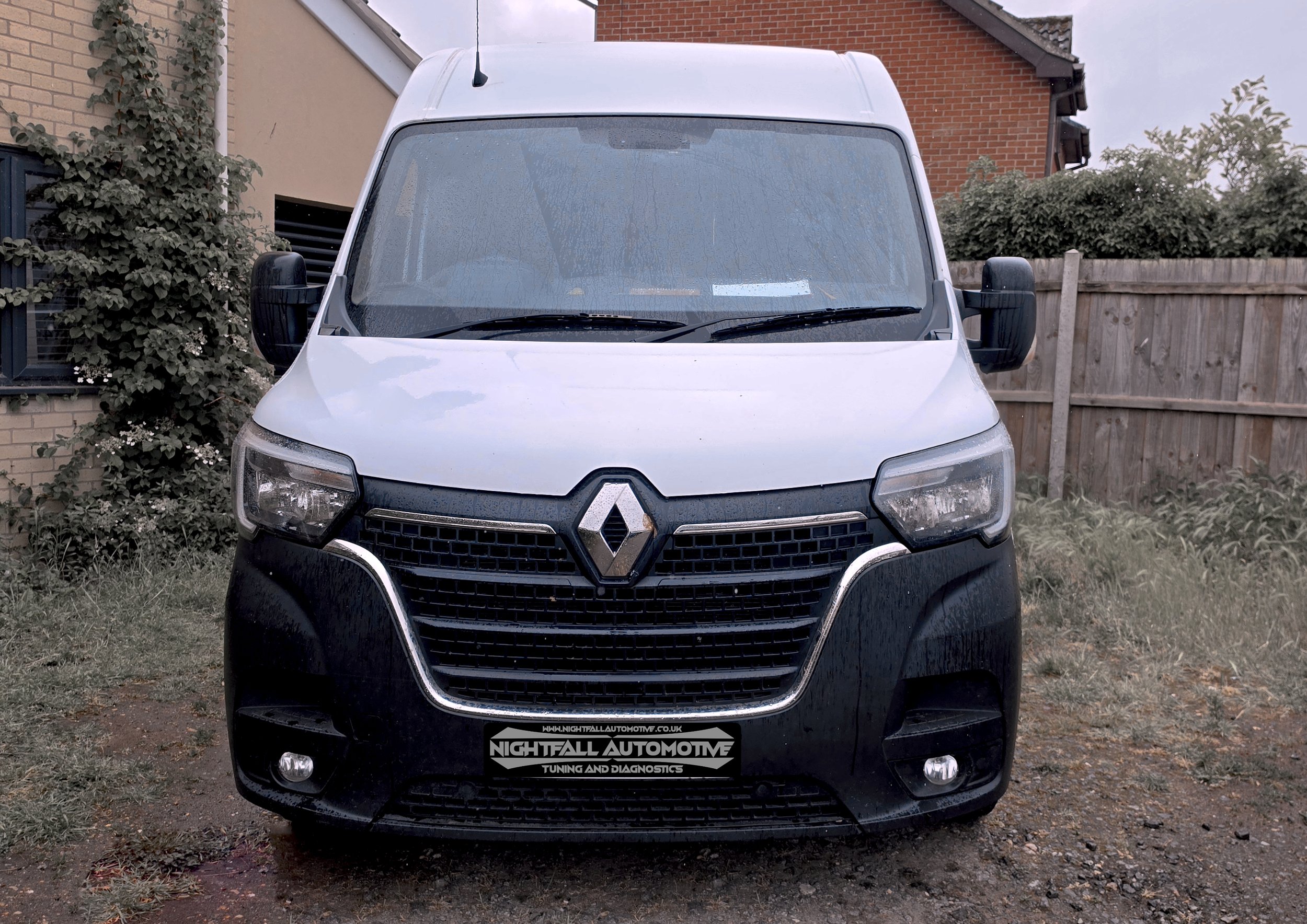 Front view of a white Renault van parked outdoors with wet windows, surrounded by a brick and wooden fence, and showing a banner for Nightfall Automotive tuning and diagnostics. tuned by Nightfall Automotive