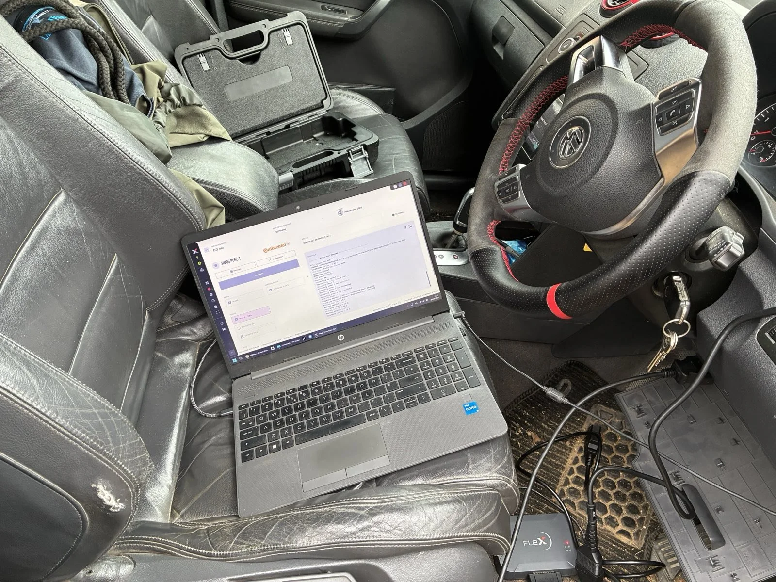 Inside a vehicle's front cabin, with a laptop on the passenger seat connected to diagnostic tools, a black steering wheel with a red stripe, and keys in the ignition.