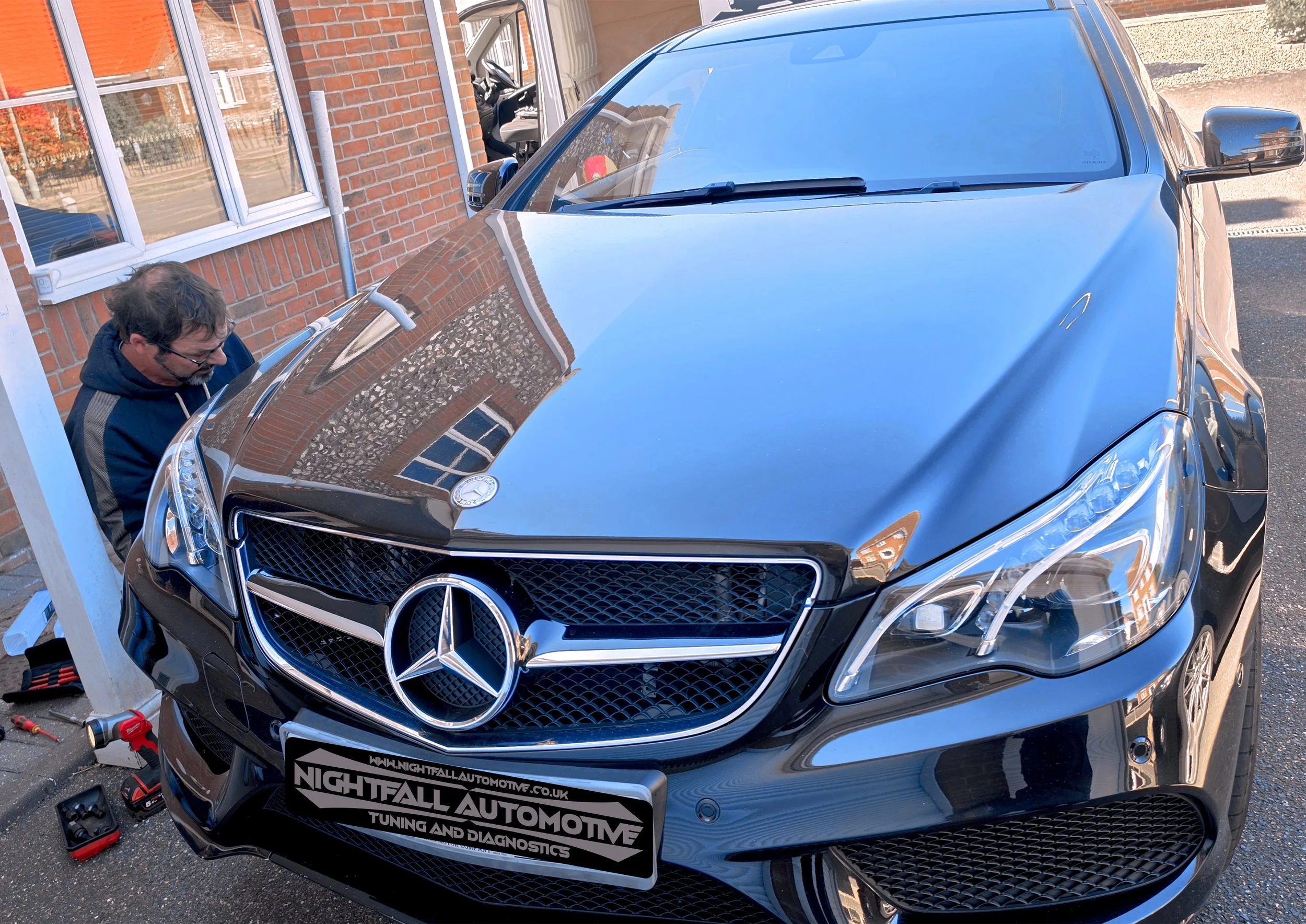 A black Mercedes-Benz car parked outside a brick building with a window. A man with glasses and a dark hoodie is working on the front of the car, with tools and a power drill nearby. The car has a sticker on the front bumper that reads 'Nightfall Aut