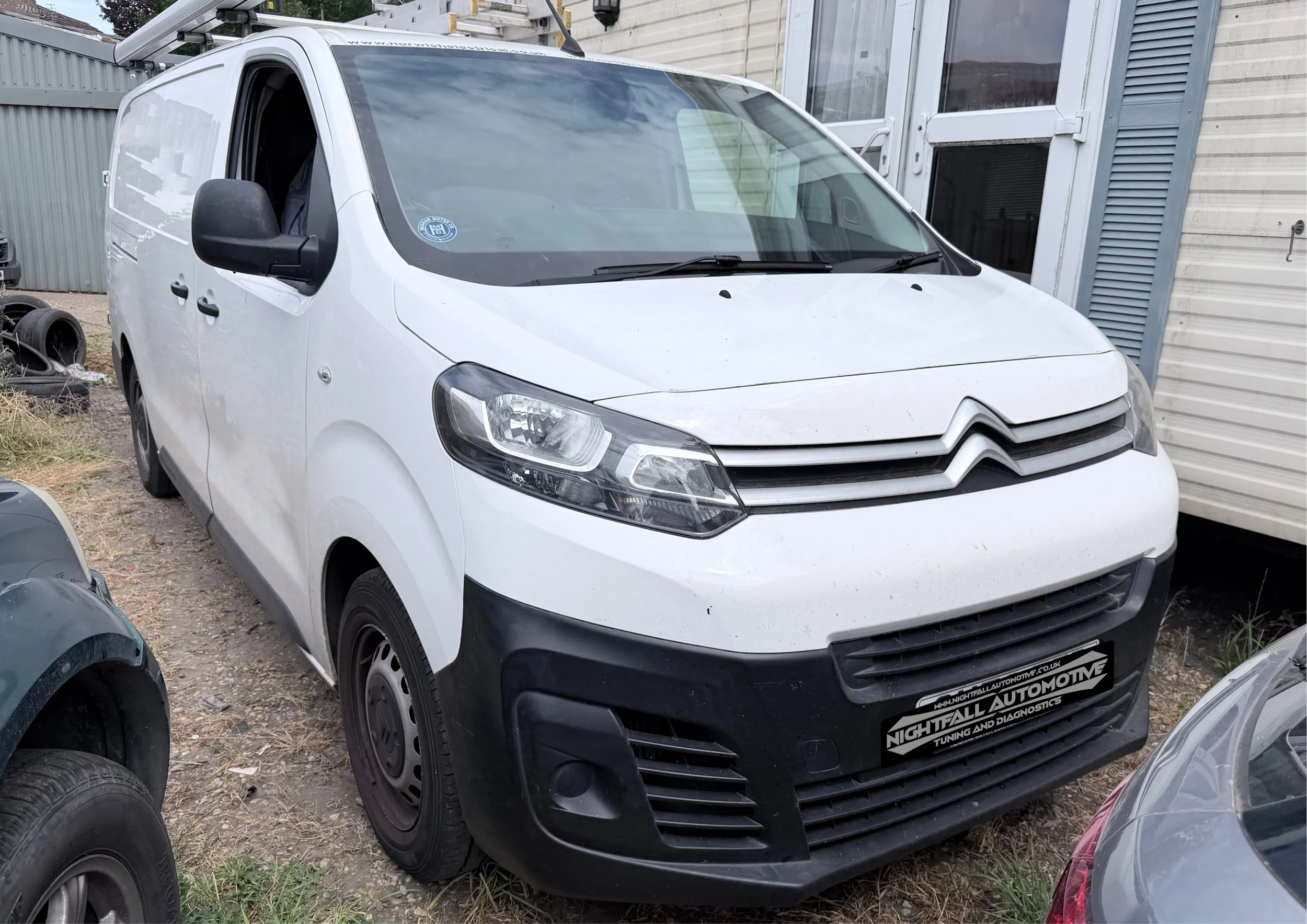 A white Citroën van parked outdoors next to a building with corrugated metal siding and a white door with a handle. The van has a black grille and headlights, and a sticker on the windshield. There are tires and car parts on the ground nearby. Tuned 