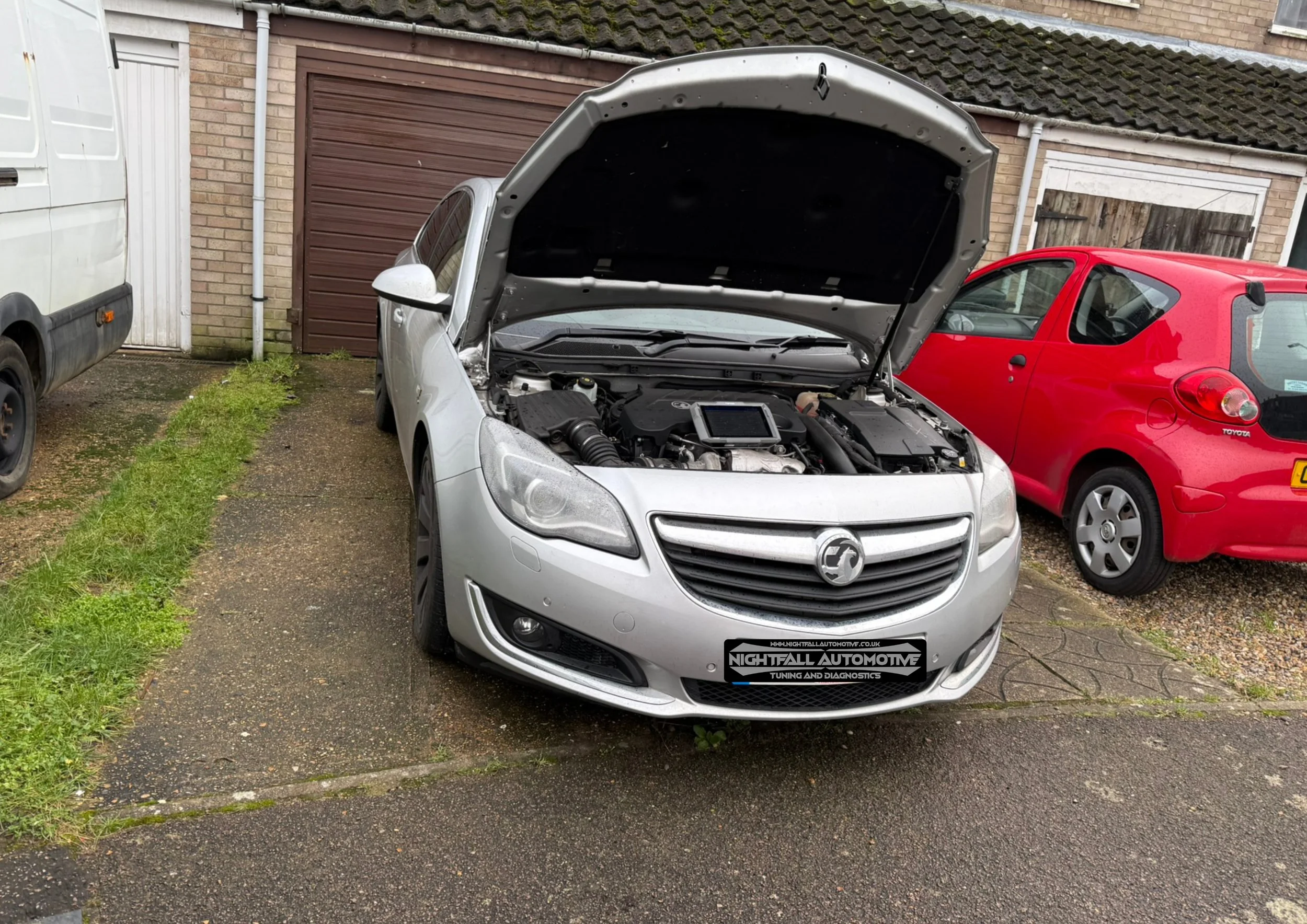 A silver hatchback car with its hood open, parked outdoors on a driveway. There are other vehicles and a brick building in the background.