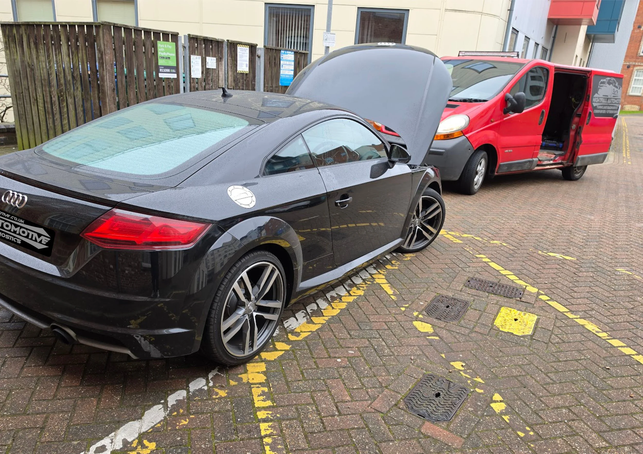 A black Audi sports car with its hood open parked next to a red service van with the side door open, in a parking lot with brick pavement and yellow parking space lines.