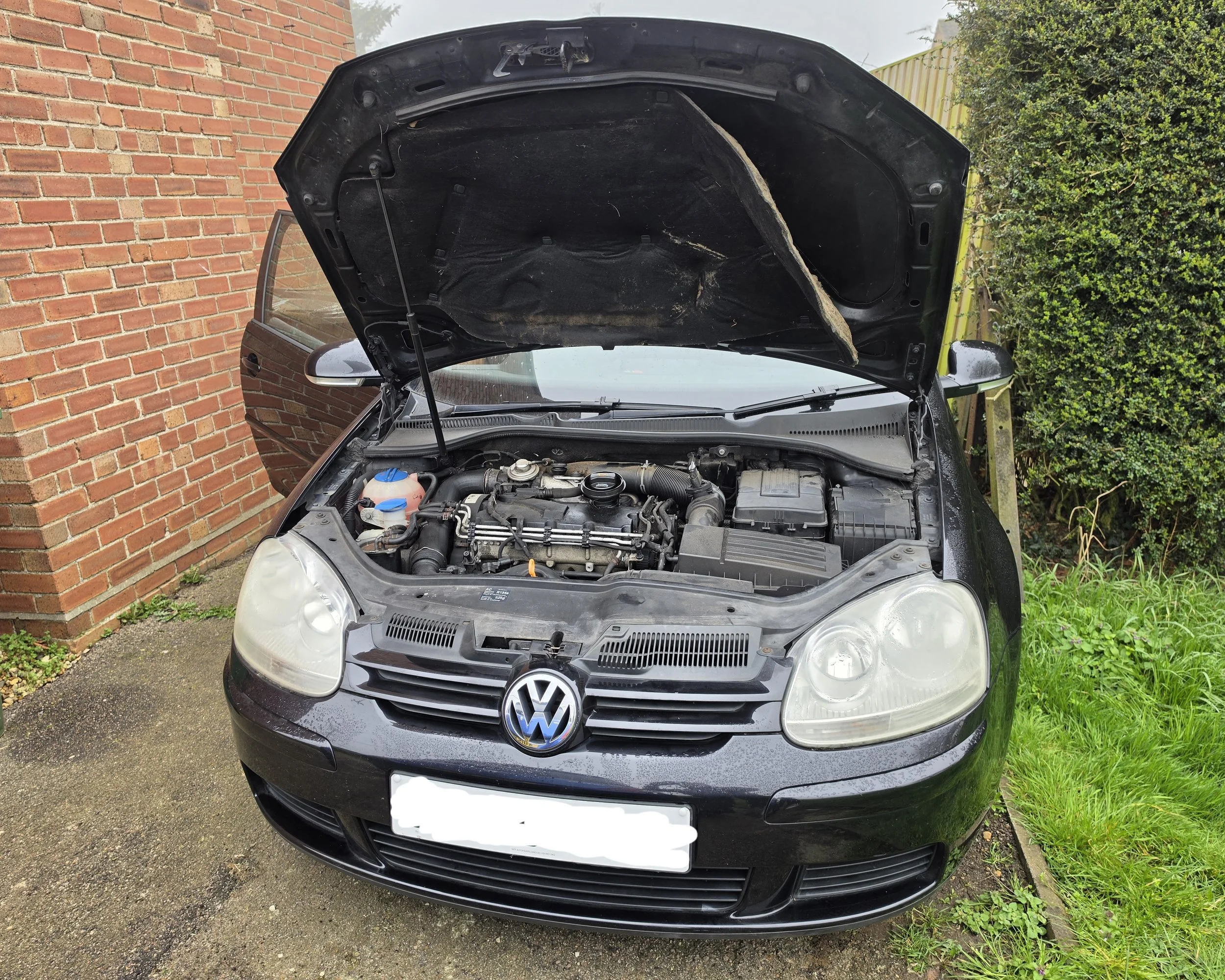 A black Volkswagen car with its hood open, parked on a driveway next to a brick wall and green bushes, showing the car's engine compartment.