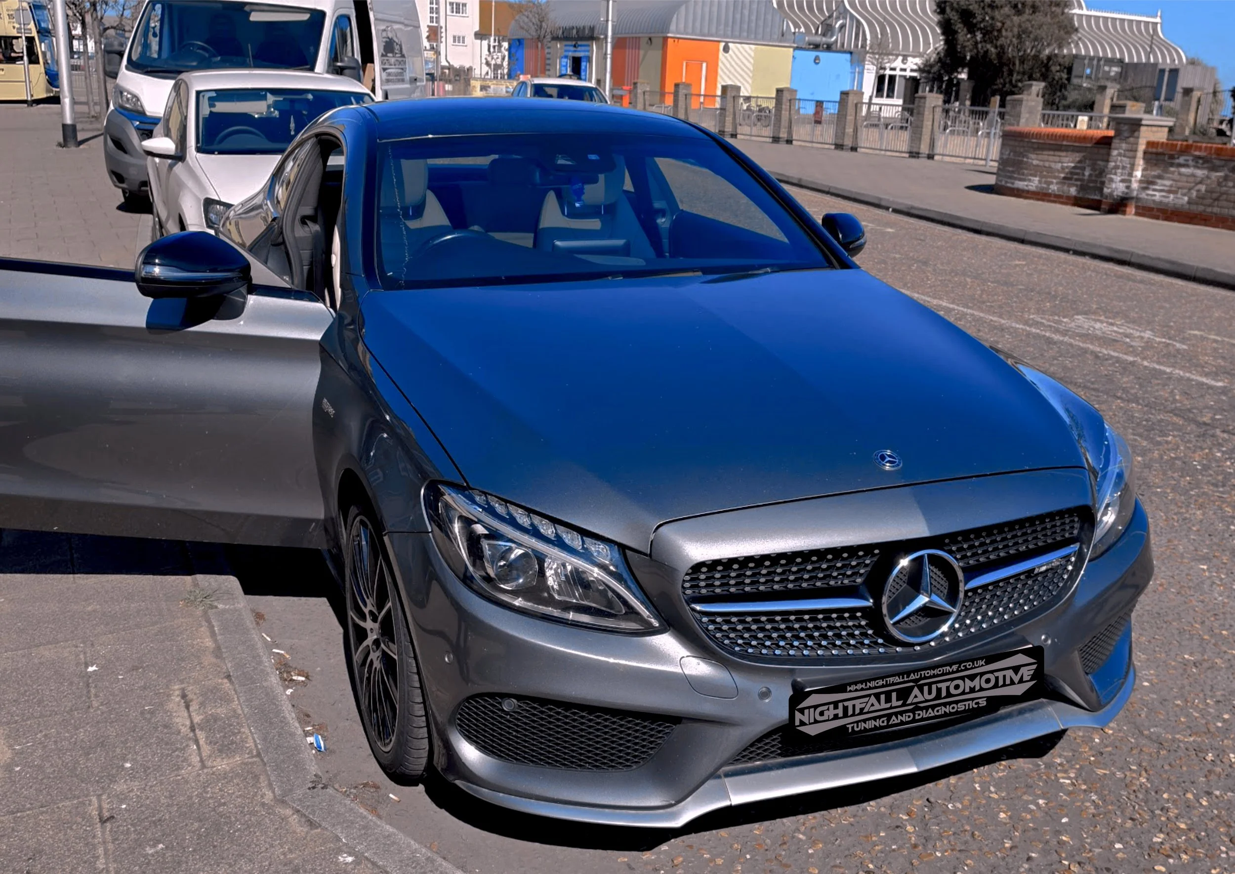A silver Mercedes-Benz parked on the street with other vehicles in the background. The car has a sign on the front bumper for Nightfall Automotive, which offers tuning and diagnostics.