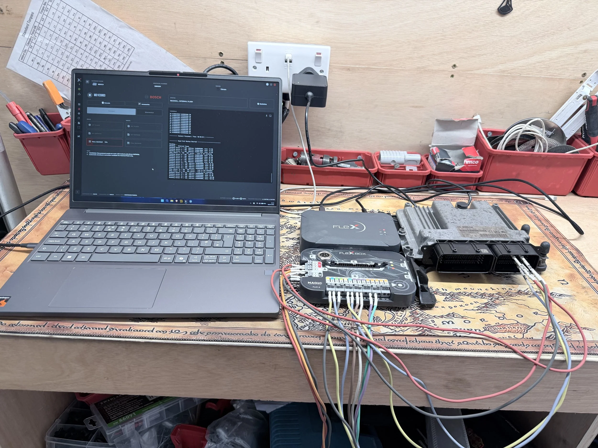 A workbench with a laptop displaying a diagnostic software screen, various tools in red containers, wiring and electronic components, and a power strip on a wooden surface. Tuned by Nightfall Automotive