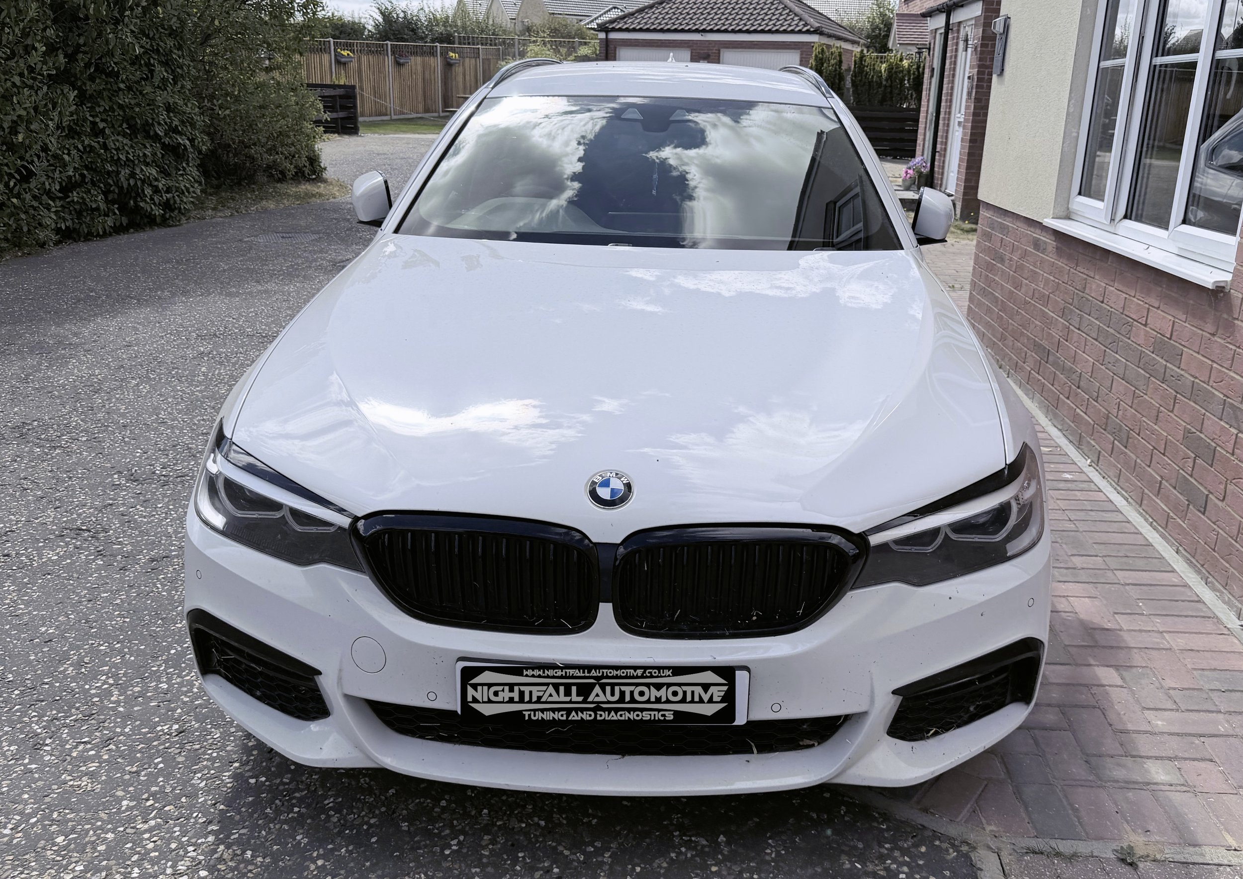 White BMW parked in a residential driveway with buildings, bushes, and a wooden fence in the background. Tuned by Nightfall Automotive