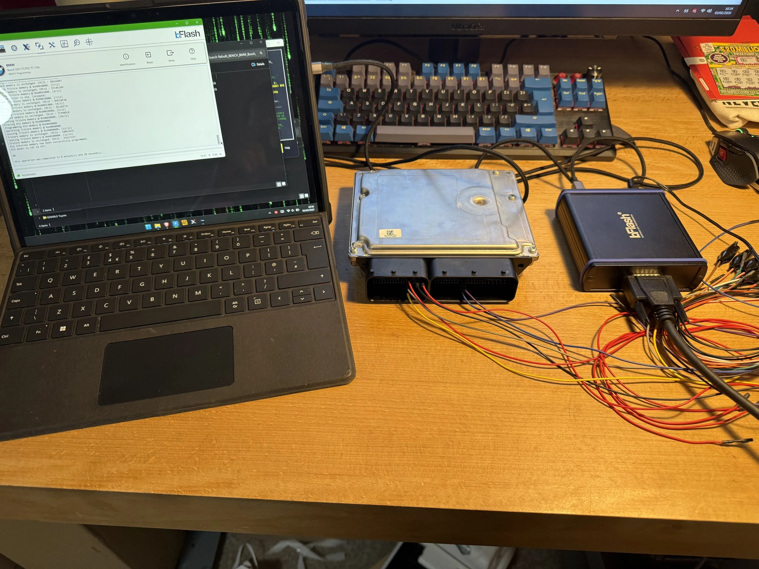 Computer repair workspace featuring a laptop, automotive engine control unit (ECU), power supply device, and wiring, on a wooden desk with a keyboard and monitor in the background.