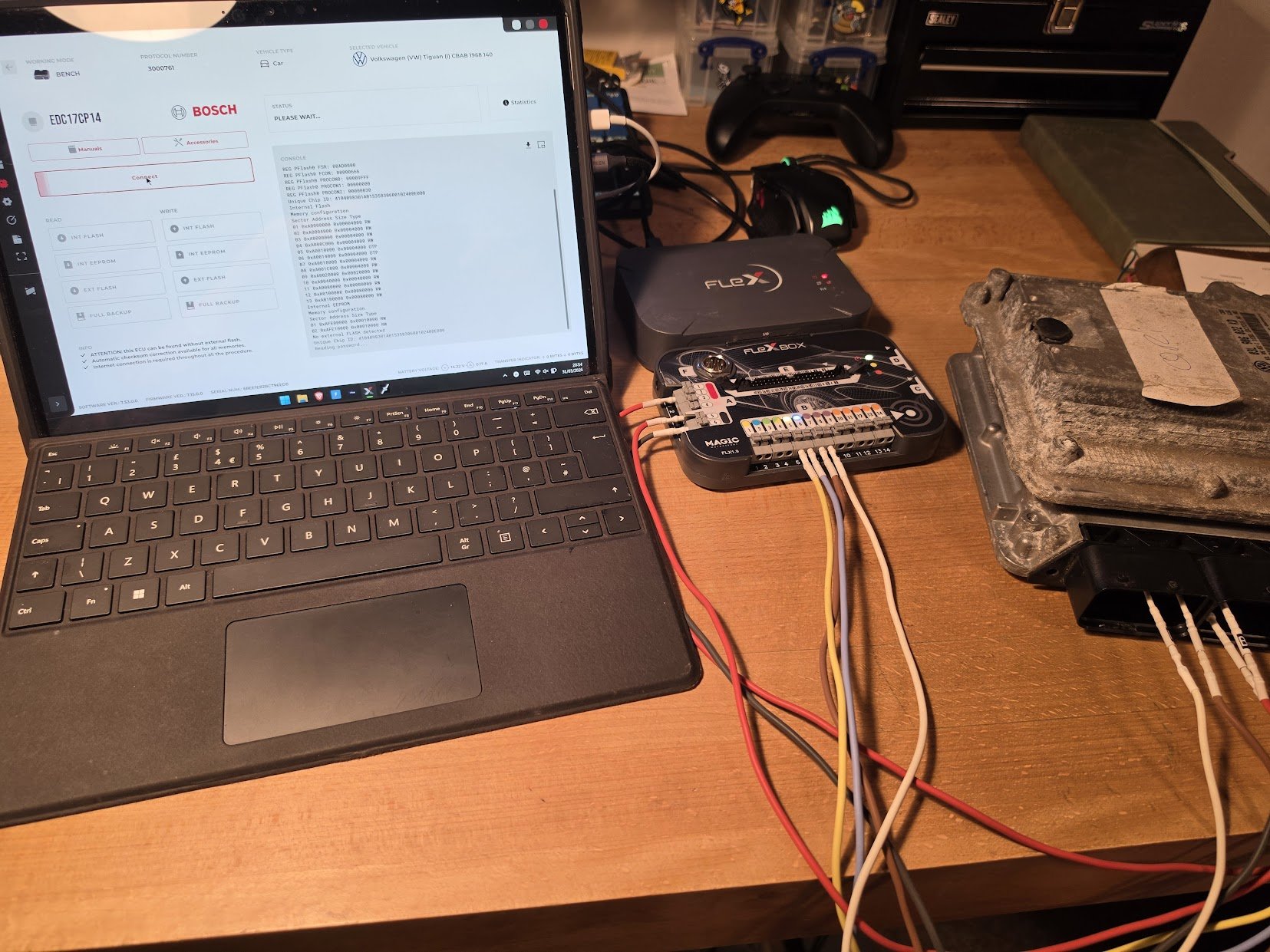 Laptop connected to a vehicle module, with wires and tools on a workshop table.