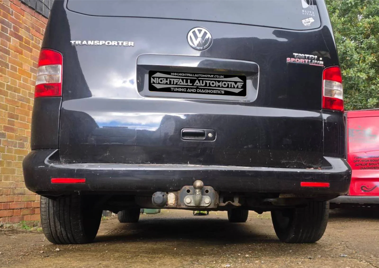 Rear view of a black Volkswagen transporter vehicles with a tow hitch and some rust, parked on a dirt surface next to a brick wall, with another red vehicle partially visible on the right.